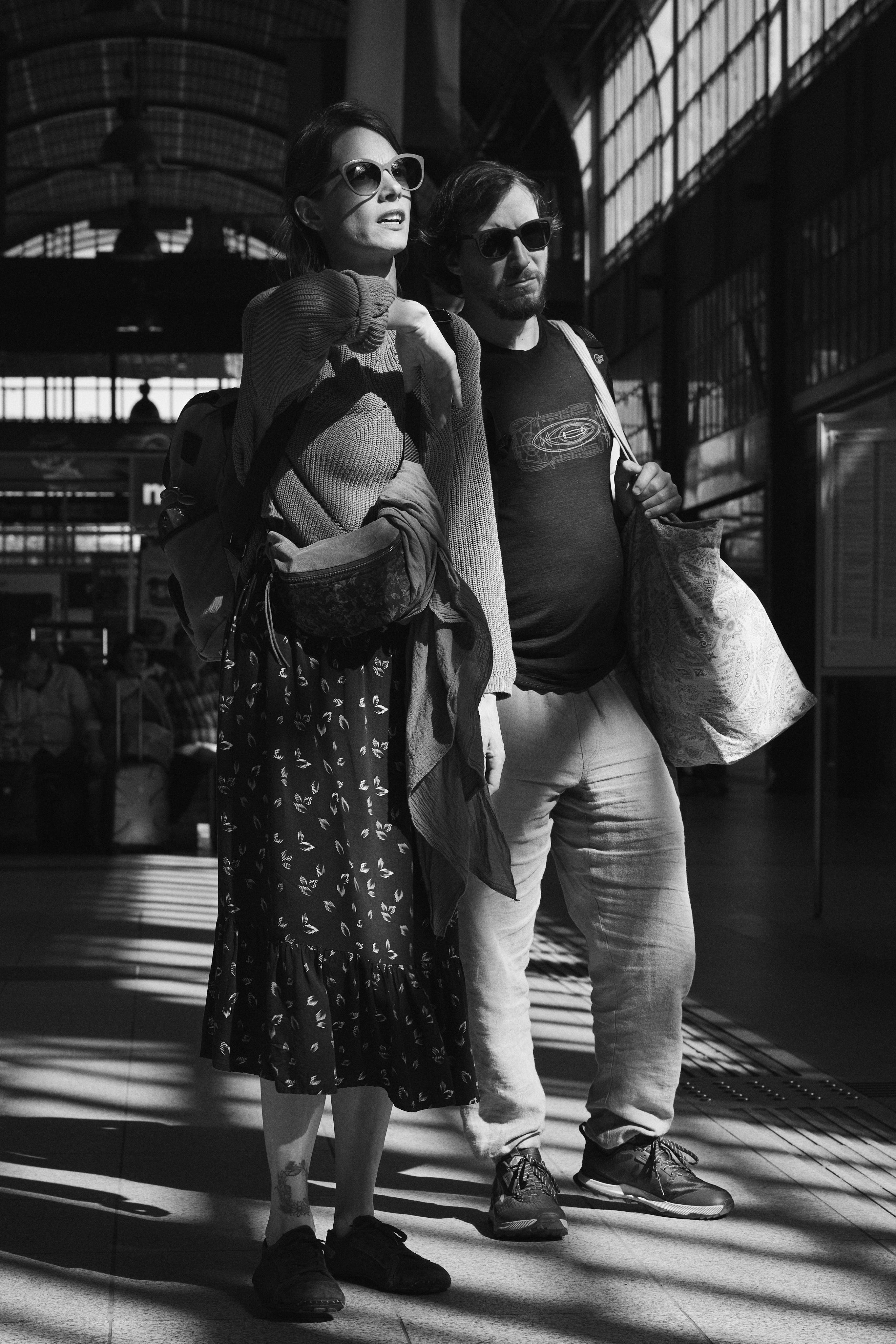 The image shows a black-and-white photograph of a man and a woman, both wearing sunglasses and seemingly engaged with each other. The woman is wearing a patterned dress and a light sweater, while the man is dressed in a casual t-shirt and pants. They both have bags slung over their shoulders and are standing inside a building with large windows, which appears to have a high ceiling. The lighting indicates strong sunlight streaming in, casting shadows and enhancing the architectural details of the building. They could be in a public place such as a train station or an airport.