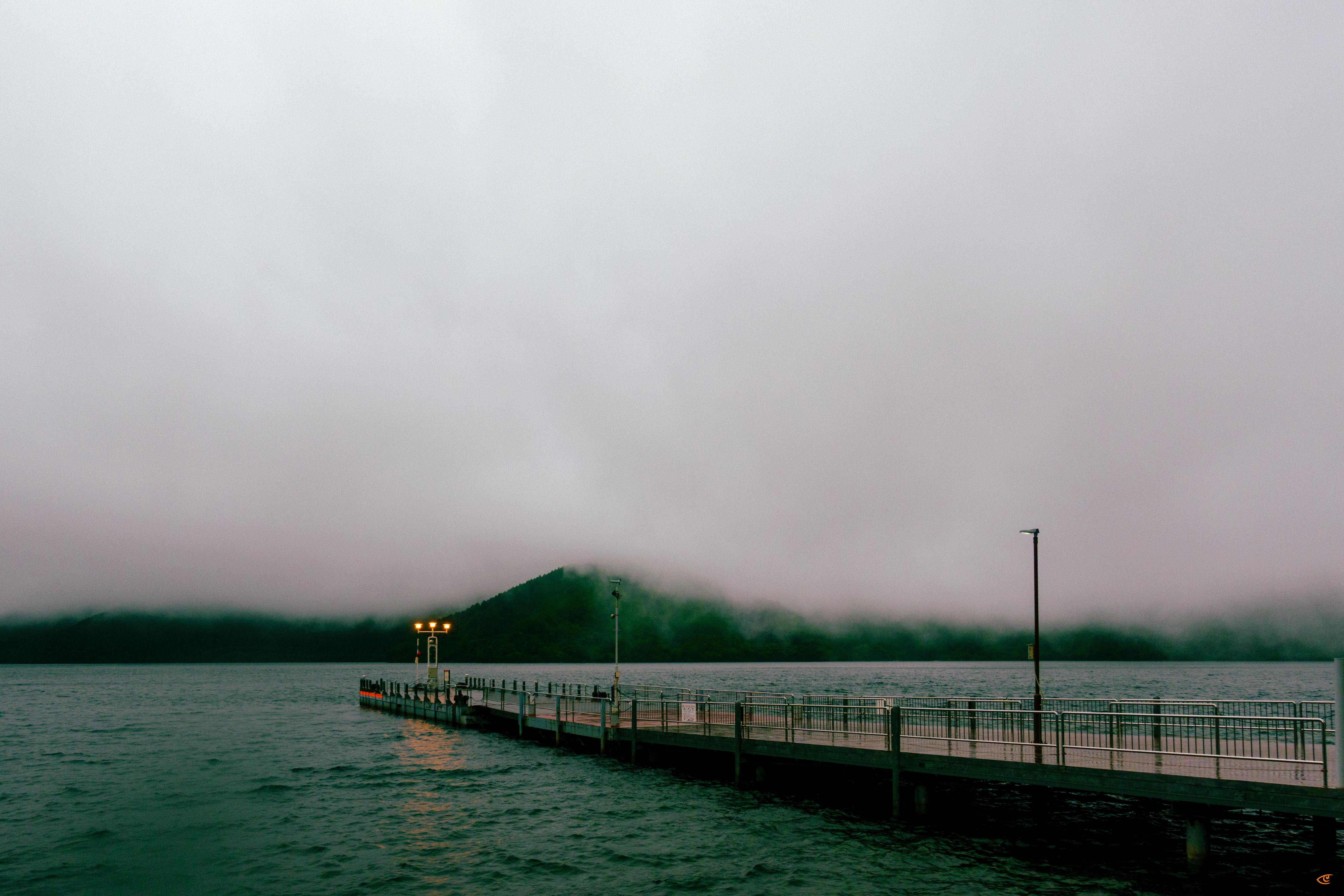 Long pier with metal railings extending into a calm lake under a gray, overcast sky, with small lights at the end and a tall lamp post along the walkway; low fog drapes over dark, tree-covered hills in the background.
