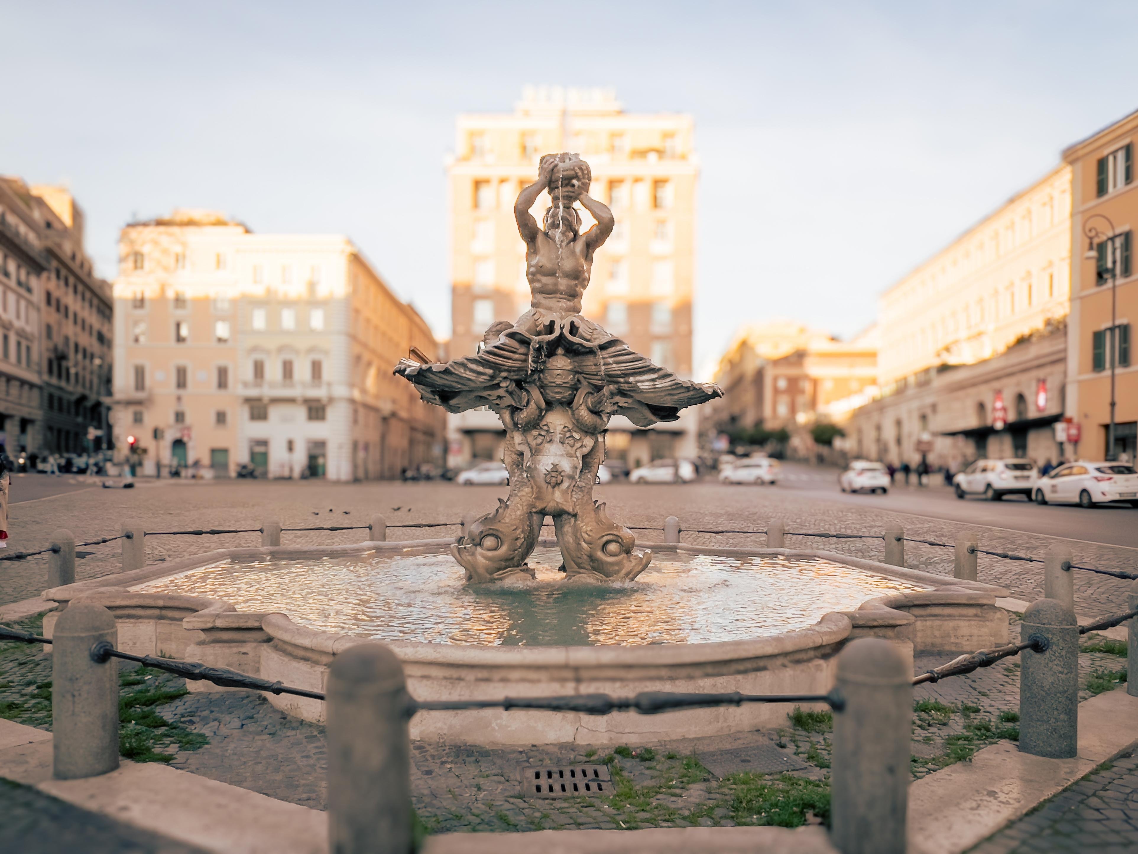 La Fontana del Tritone, una famosa fontana situata a Roma, Italia. Presenta una scultura di Tritone, un dio del mare della mitologia greca, raffigurato mentre tiene una conchiglia. La fontana si trova in un contesto urbano con edifici sullo sfondo.