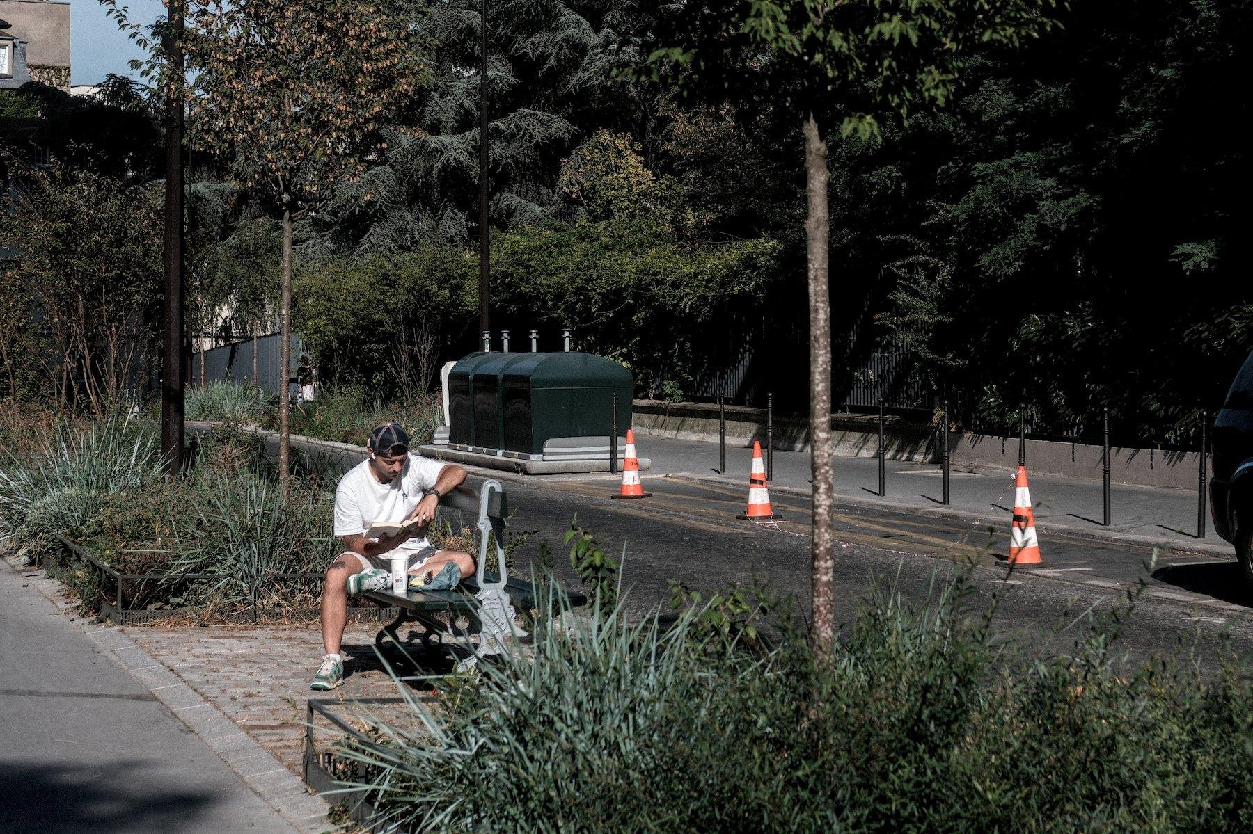 Photo horizontale. Sur un banc publique parisien vert double, un homme est assis accoudé au dossier, lisant un livre, casquette portée à l’envers, t-shirt et short blancs