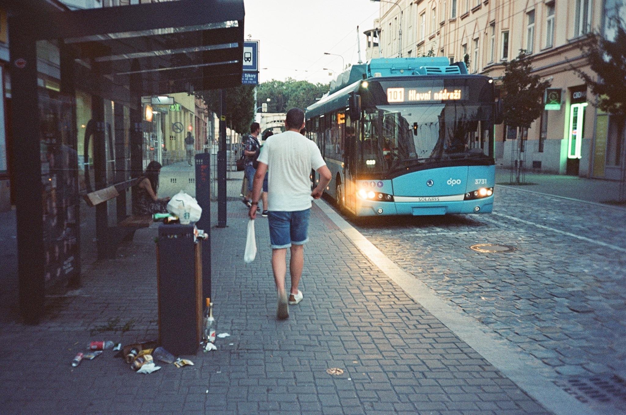 Man walking along a sidewalk beside a bus stop with scattered litter near a trash bin, as a blue city bus approaches on a cobblestone street lined with buildings.