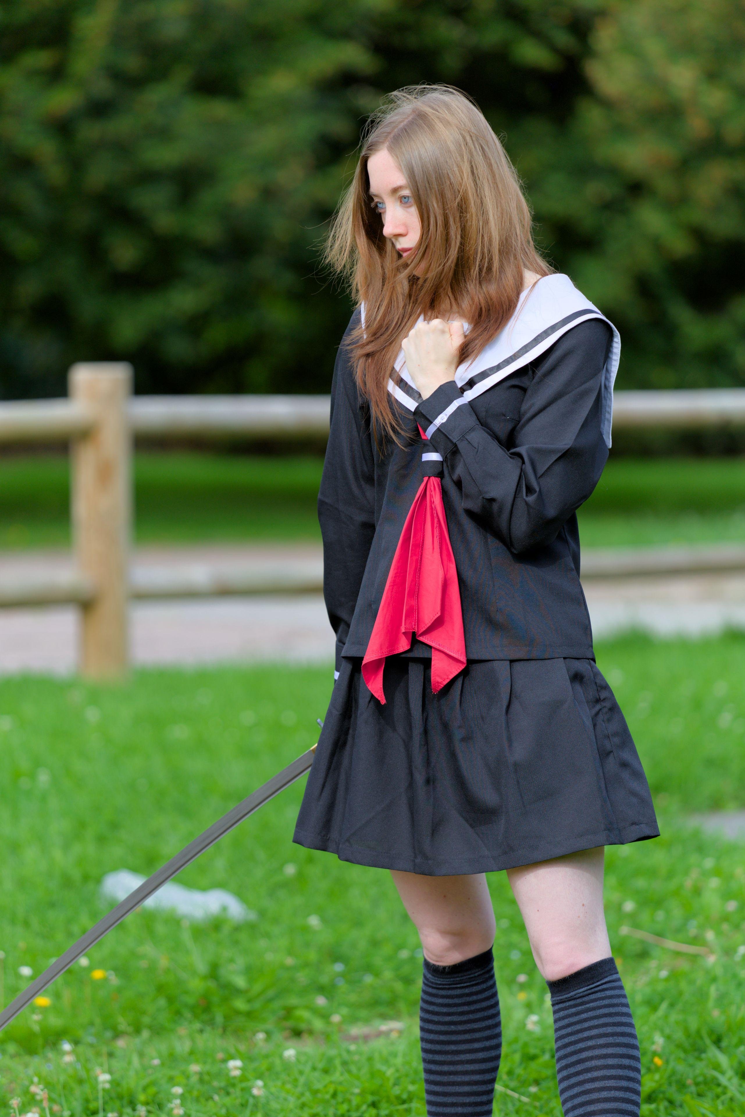 Photo format portrait d’une jeune femme rousse, cheveux long légèrement poussés vers l’avant par le vent, en uniforme d’écolière japonais bleu marine avec un ruban rouge et col blanc, tenant un katana de la main droite, bras tendu vers le bas, main cachée par la jupe. La main gauche serre le poing, tenu sur le sternum, les yeux acier regardant devant elle