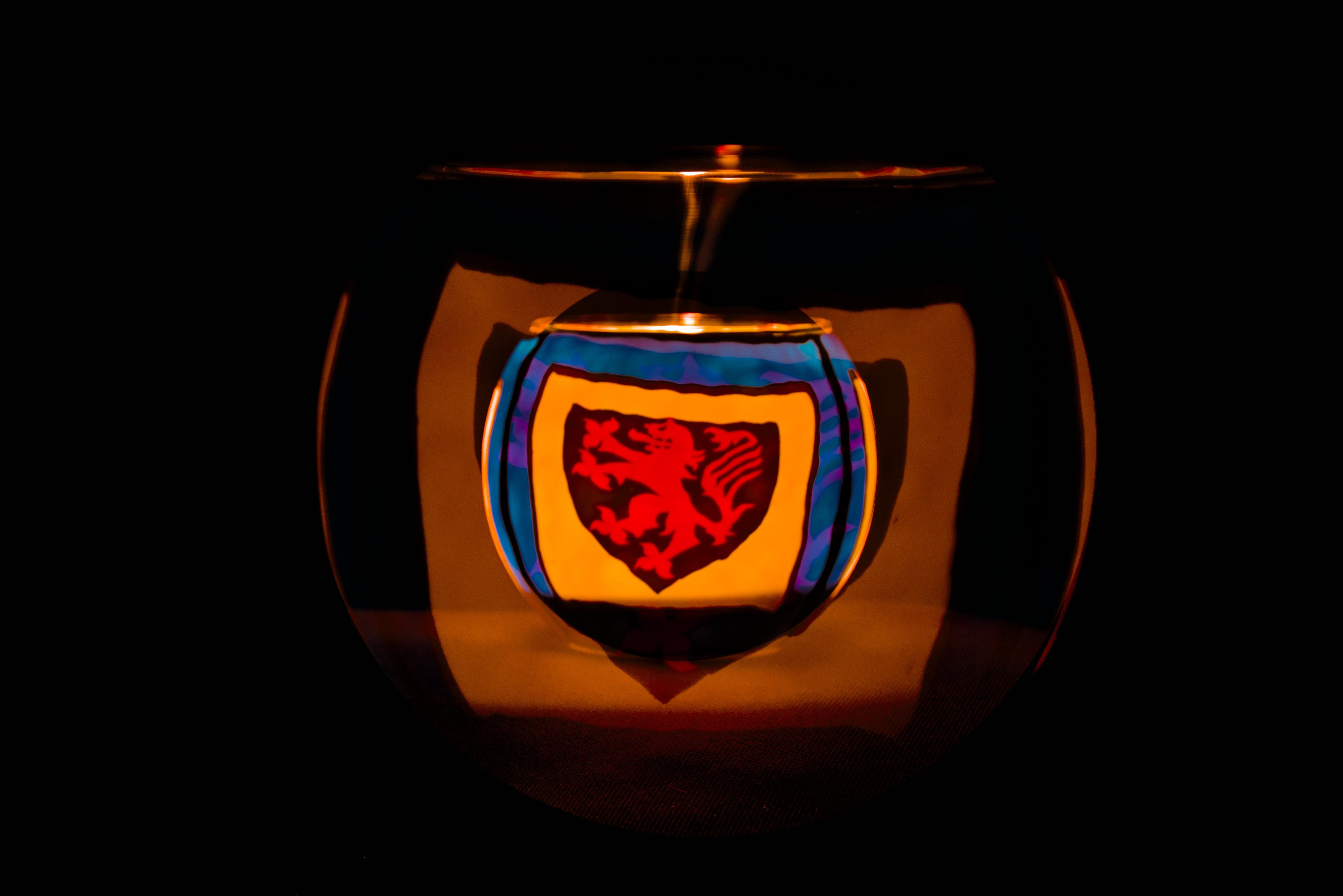 Das Bild zeigt ein Glaskugel mit einer Kerze im Inneren mit einem heraldischen Emblem, das einen roten Löwen auf kontrastierendem Hintergrund zeigt. Die Beleuchtung erzeugt einen reflektierenden Effekt, der das Emblem und seine Farben hervorhebt. Der Hintergrund ist dunkel und hebt das zentrale Design hervor.
###
The image shows a glass globe with a candle inside with a heraldic emblem depicting a red lion on a contrasting background. The lighting creates a reflective effect that highlights the emblem and its colours. The background is dark, highlighting the central design.