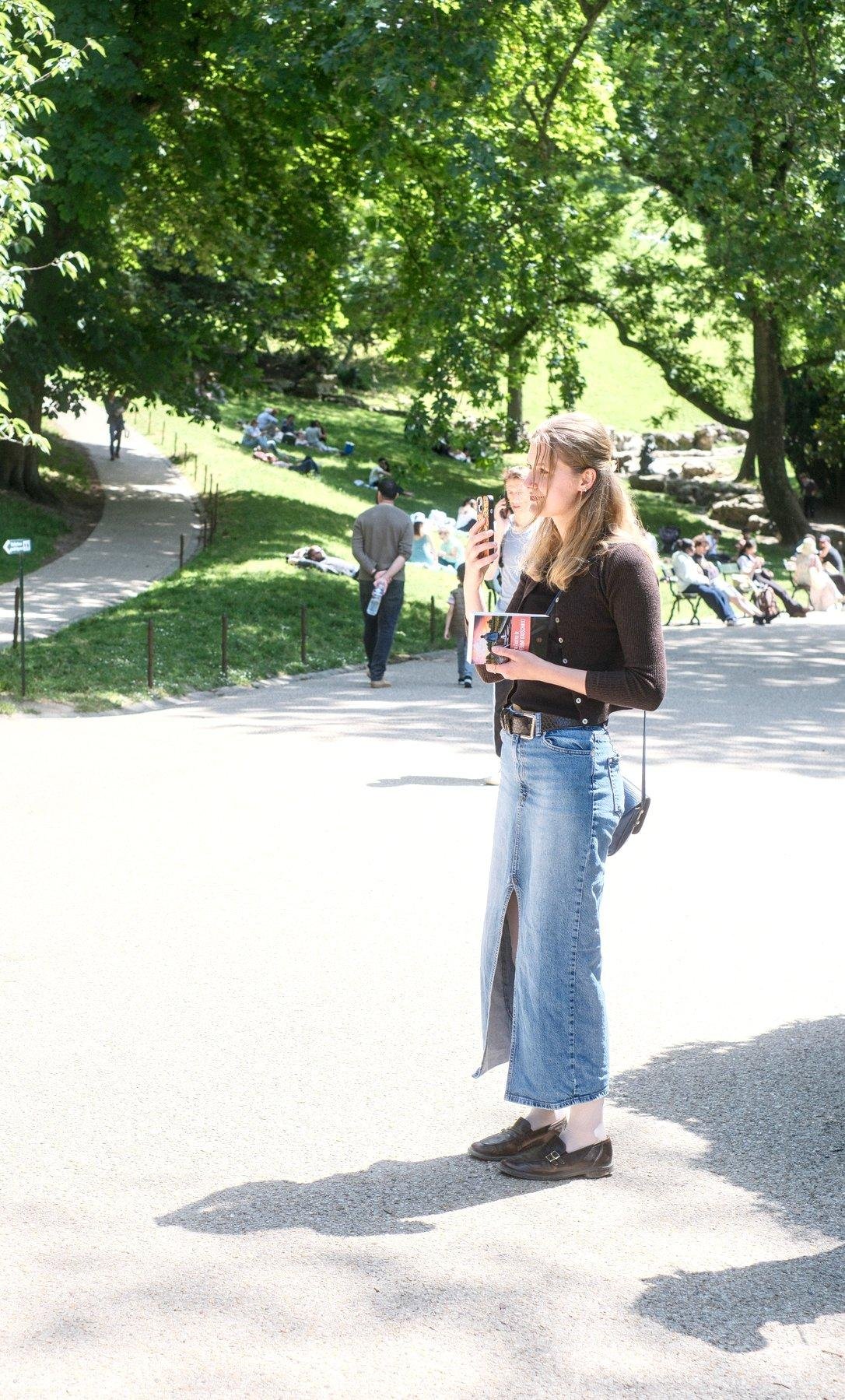 Photo verticale. Une jeune femme en longue jupe en denim, chaussure noire, gilet marron, cheveux châtains clairs, portant un livre de la main gauche et filmant avec son téléphone de la main droite