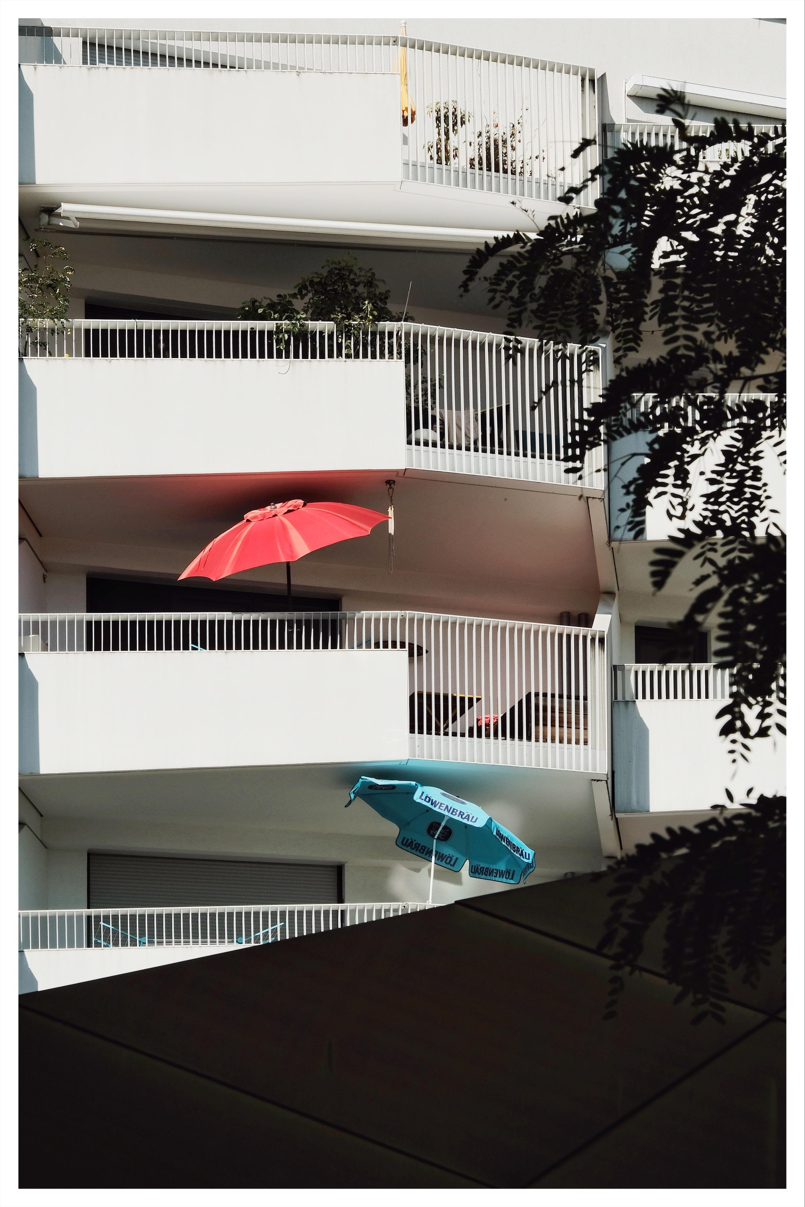 Multi-story apartment building with white balconies and railings, a red patio umbrella on a middle balcony, a blue umbrella below, and silhouetted tree branches framing the right side.