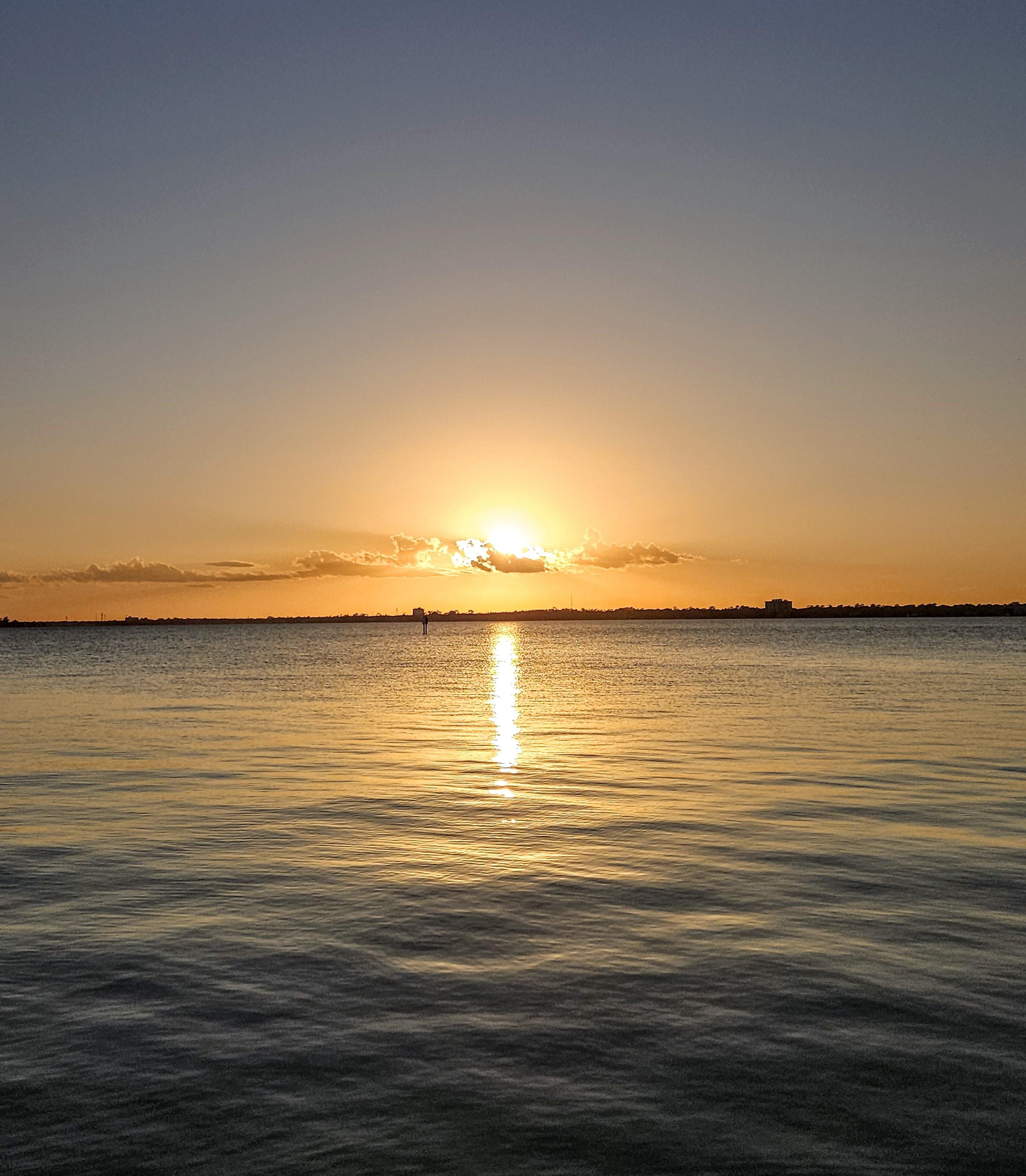 Sunset over a calm body of water, with the sun near the horizon casting a warm golden reflection across the surface. Light clouds are scattered in the sky, and a distant shoreline is visible with silhouetted structures and foliage.
