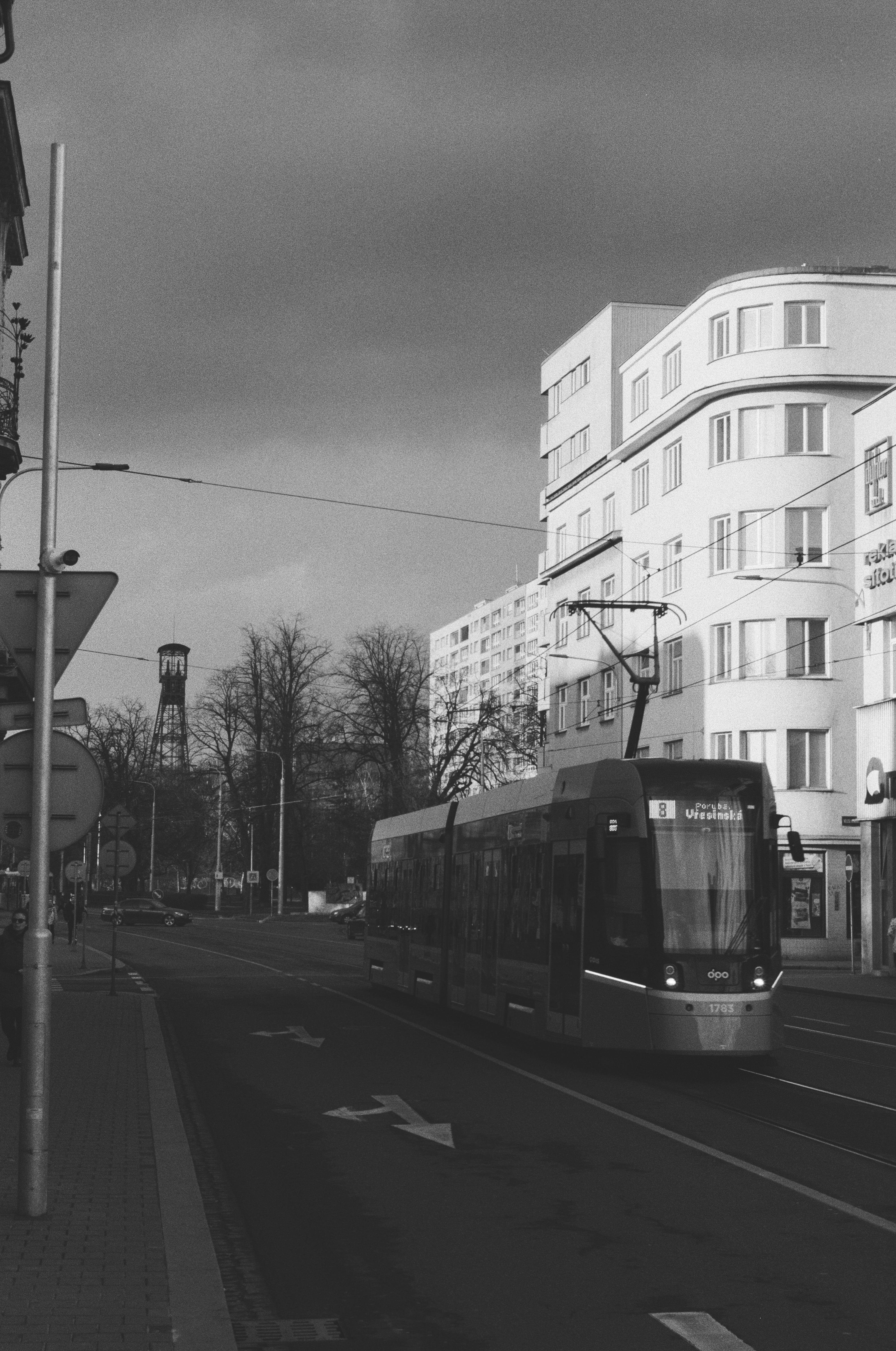A monochrome photograph of an urban street with a darkened sky, lit building, sa tram and a mining tower in the background