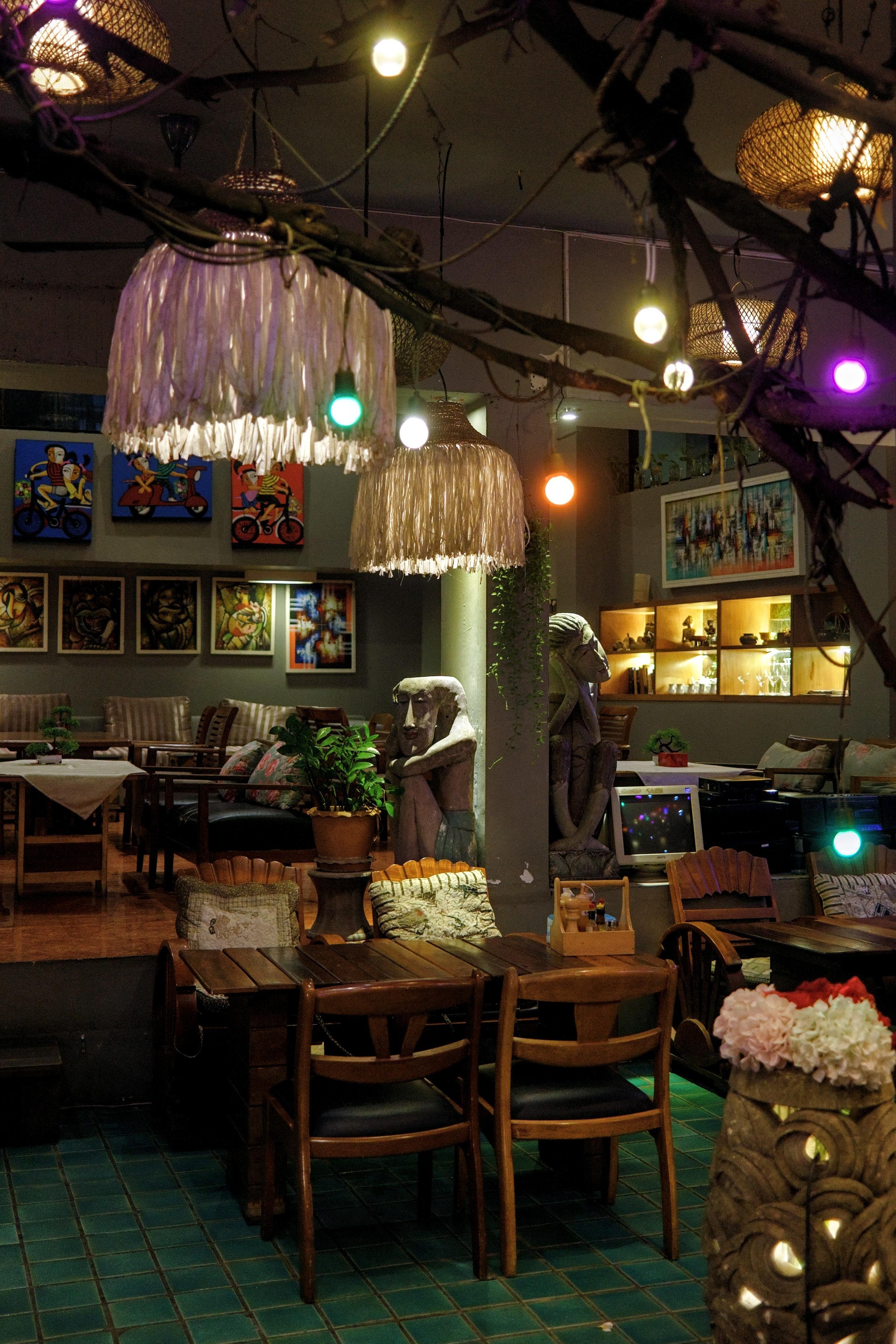 Cozy café interior with wooden tables and chairs on teal tile flooring, colorful string lights and woven pendant lamps hanging from branches overhead, framed artwork on the back wall, two carved statues near a central column, and a lit display shelf behind a seating area.