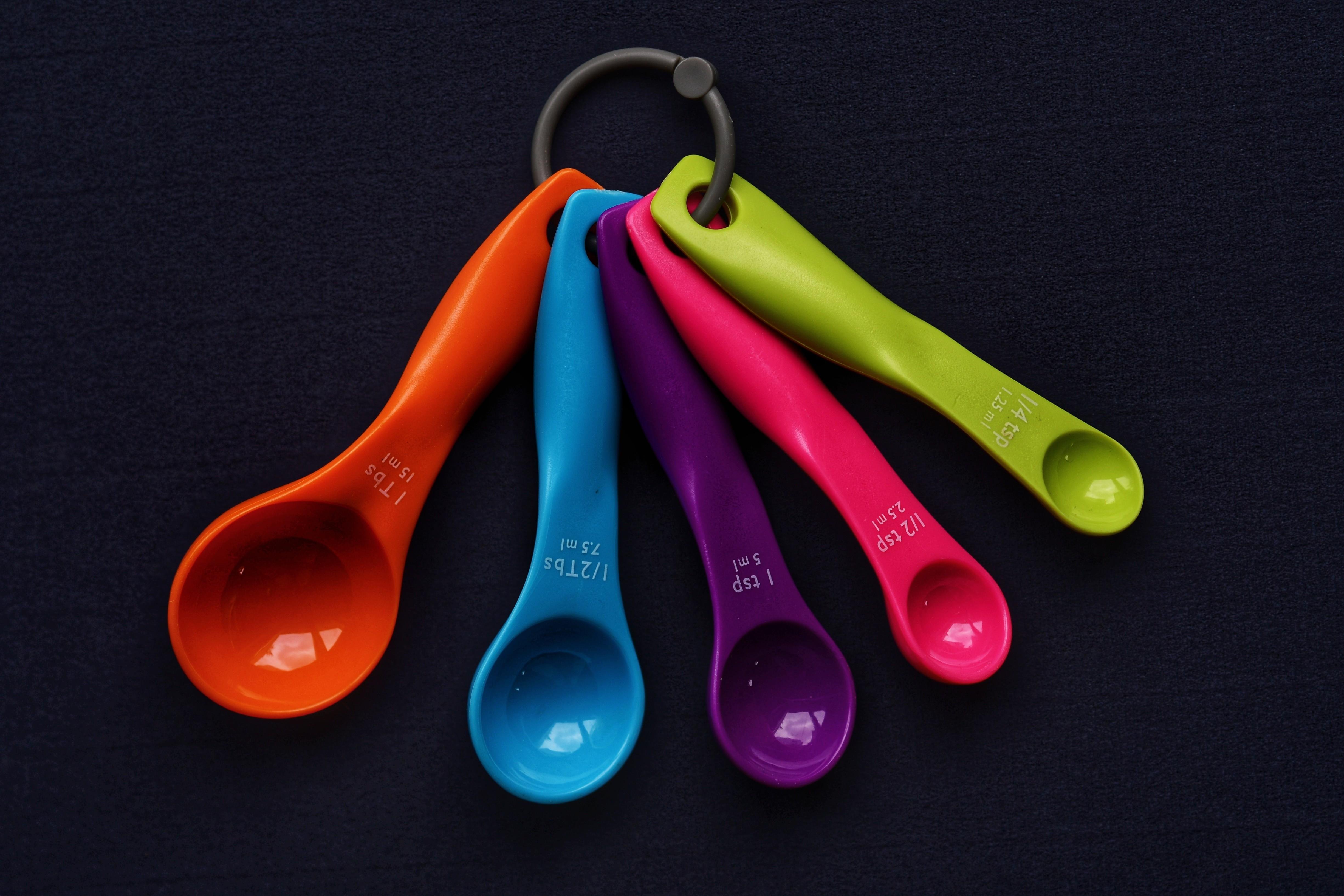 Five colorful plastic measuring spoons (orange, blue, purple, pink, and green) on a metal ring, fanned out on a dark fabric background. ### 
Fünf bunte Messlöffel aus Plastik (orange, blau, lila, rosa und grün) auf einem Metallring, fächerförmig angeordnet auf einem dunklen Stoffhintergrund.