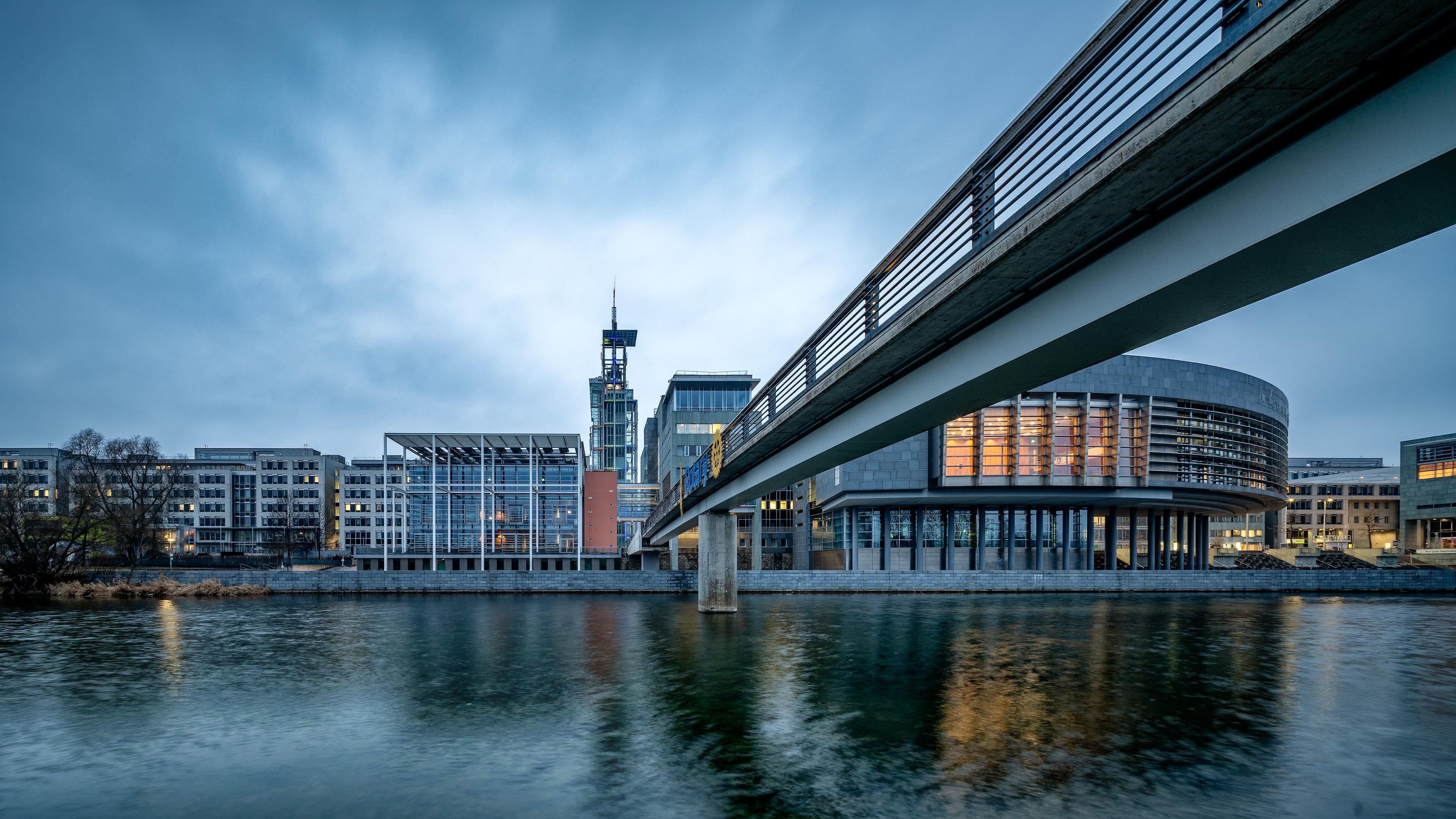 Modern city waterfront at dusk with a long pedestrian bridge crossing from the right over calm water, reflections on the surface, and illuminated glass-and-concrete buildings including a cylindrical structure and a tall tower under a cloudy sky.