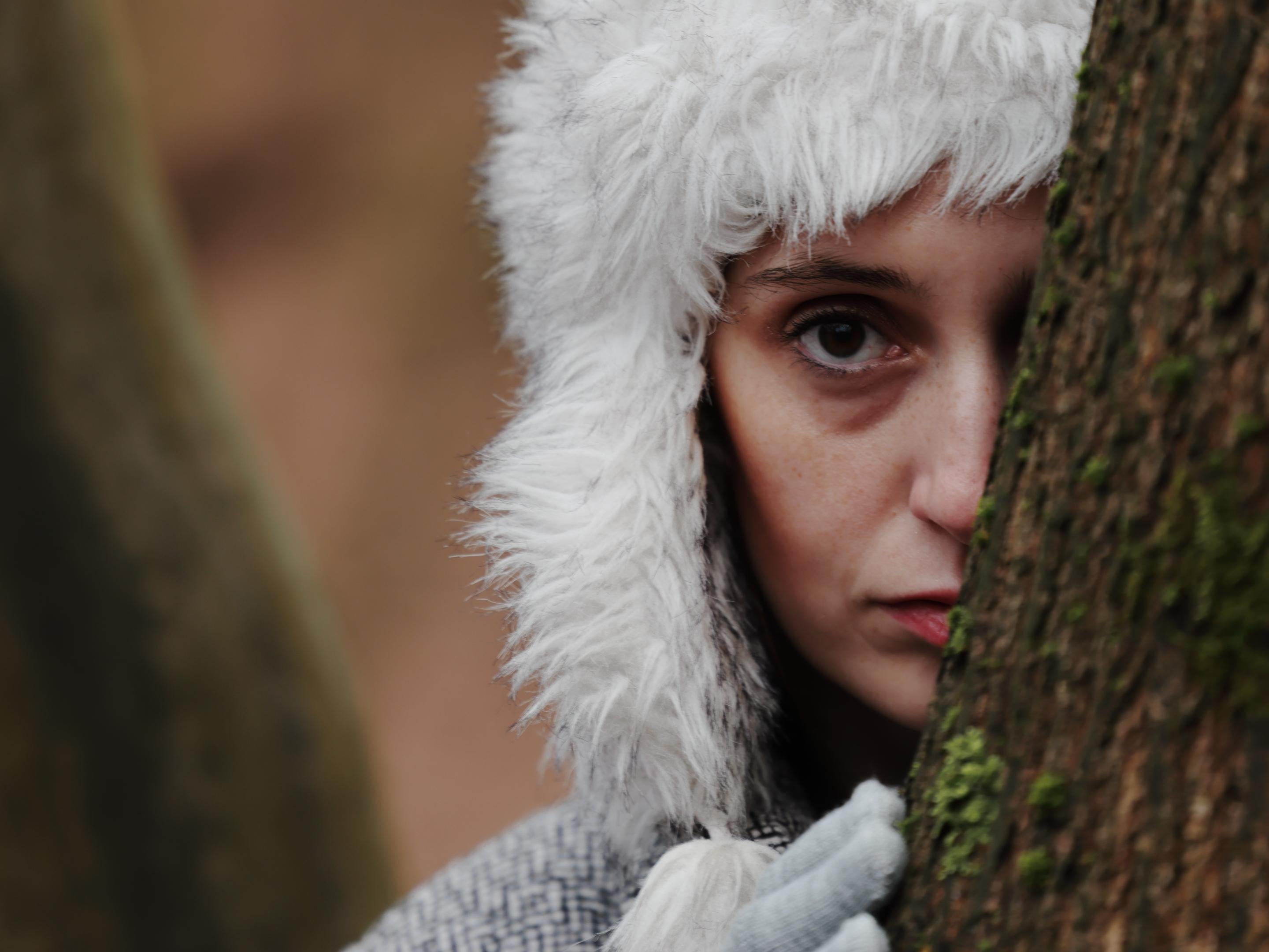 The image shows a person wearing a white, fluffy hat and looking at the camera while partially hidden behind a tree. The person has a serious expression, with one eye visible and holding the tree with their hand. The background is blurred, focusing on the person.