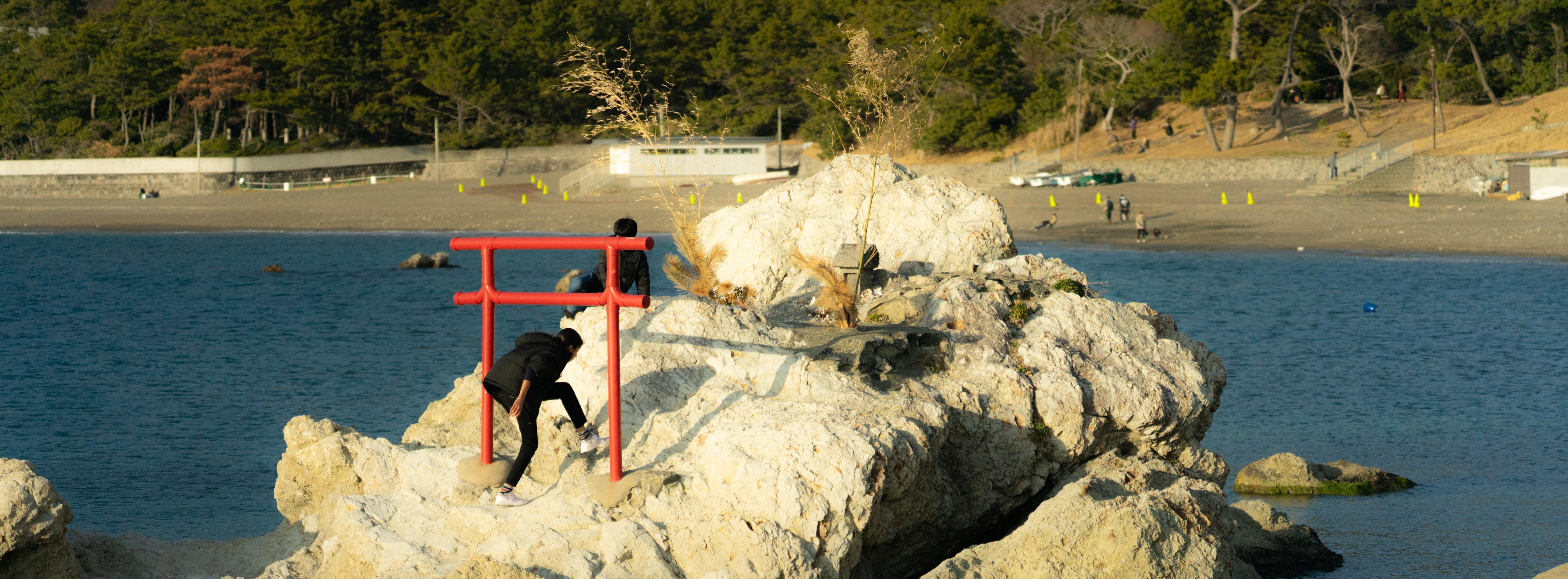 A person climbs over large, white rocks near the sea, with a red torii gate on the rocks. In the background, a sandy beach and dense, green forest line the coast, with several people scattered along the shore.
