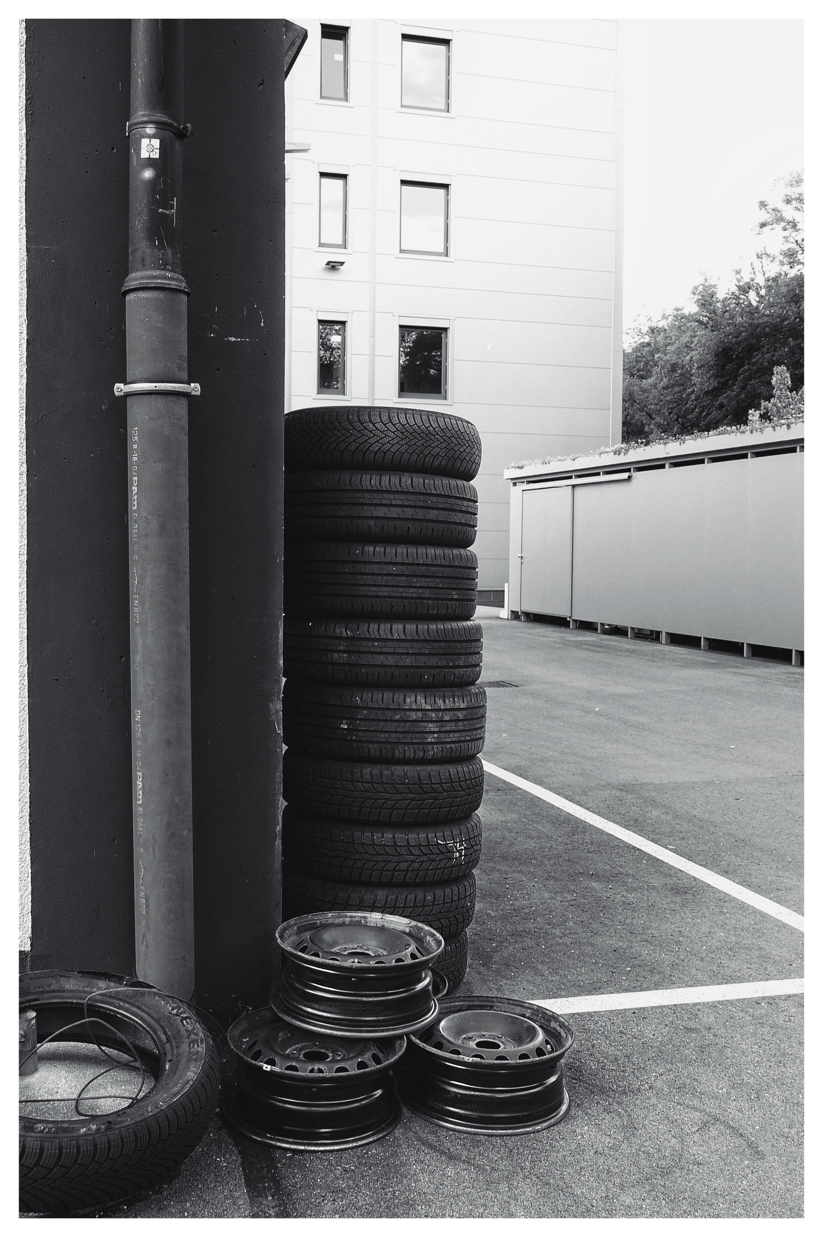 A stack of tires is leaning against a building with two metal pipes. Three tire rims are placed on the ground beside the stack. The building in the background has several windows and corrugated metal siding. The scene is set in a parking lot with visible parking lines on the pavement. The image is in black and white.