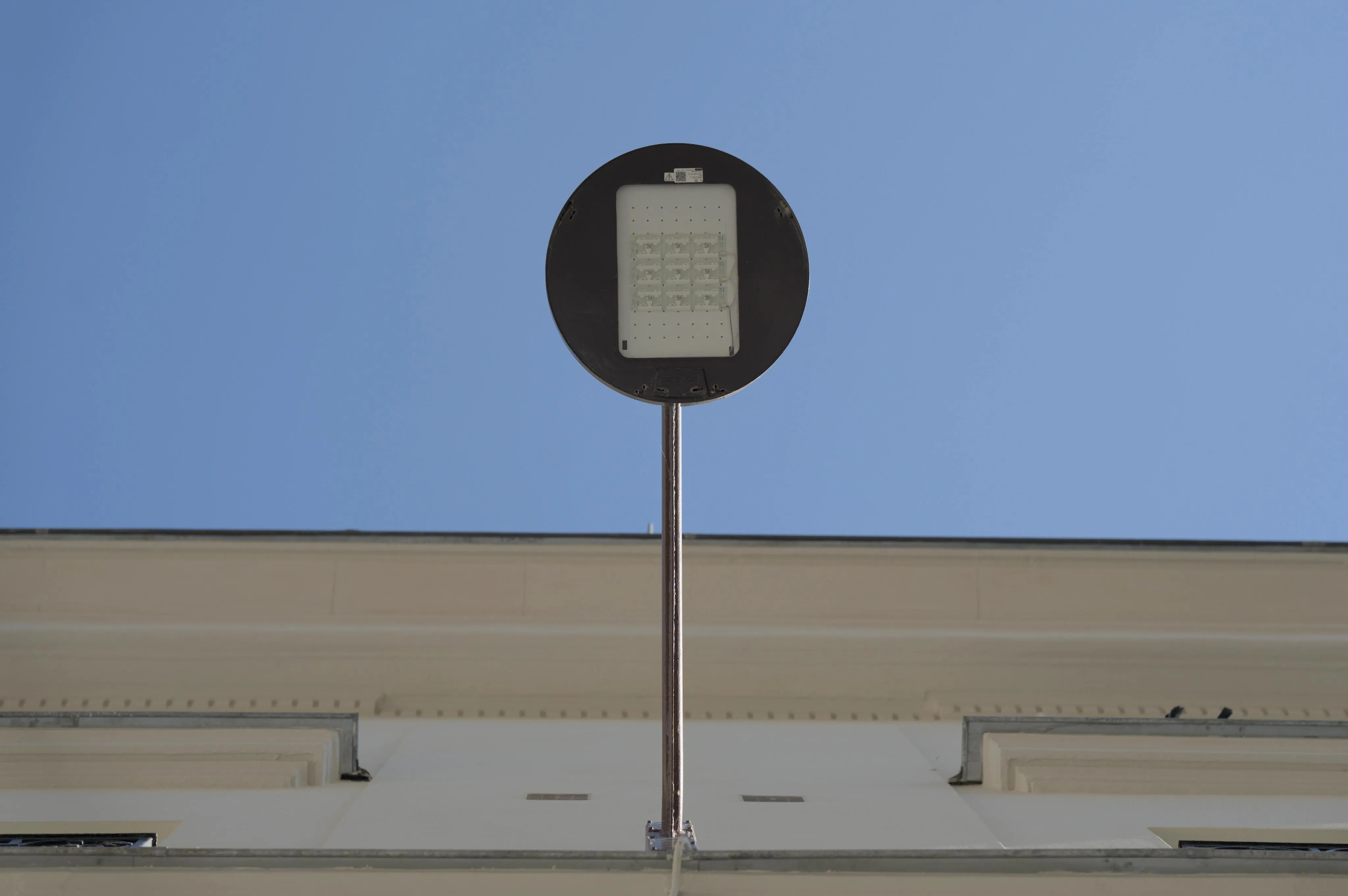 Photograph of a round street lamp hanging from the side of a building, seen from vertically underneath. The vertical perspective makes the image look like the lamp is a round sign at the top end of a pole. The side of the building is smooth and painted white. In the background, the sky is blue and cloudless.