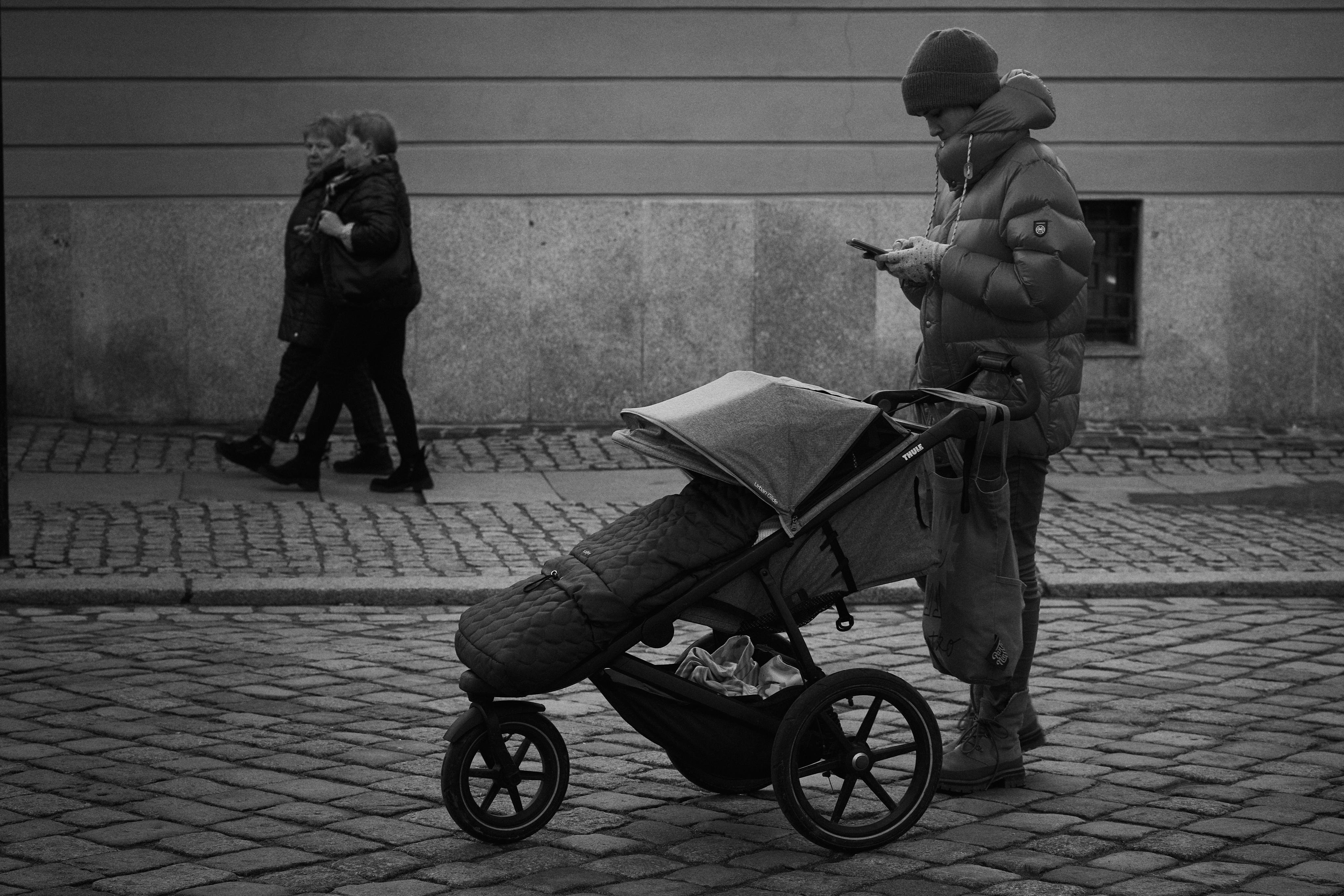 The image is a black and white photo of a street scene. It shows a person wearing a winter jacket and hat, looking at their phone while standing beside a stroller on a cobblestone road. In the background, there are two people walking and talking. The setting appears to be in an urban area with a building wall in the background.