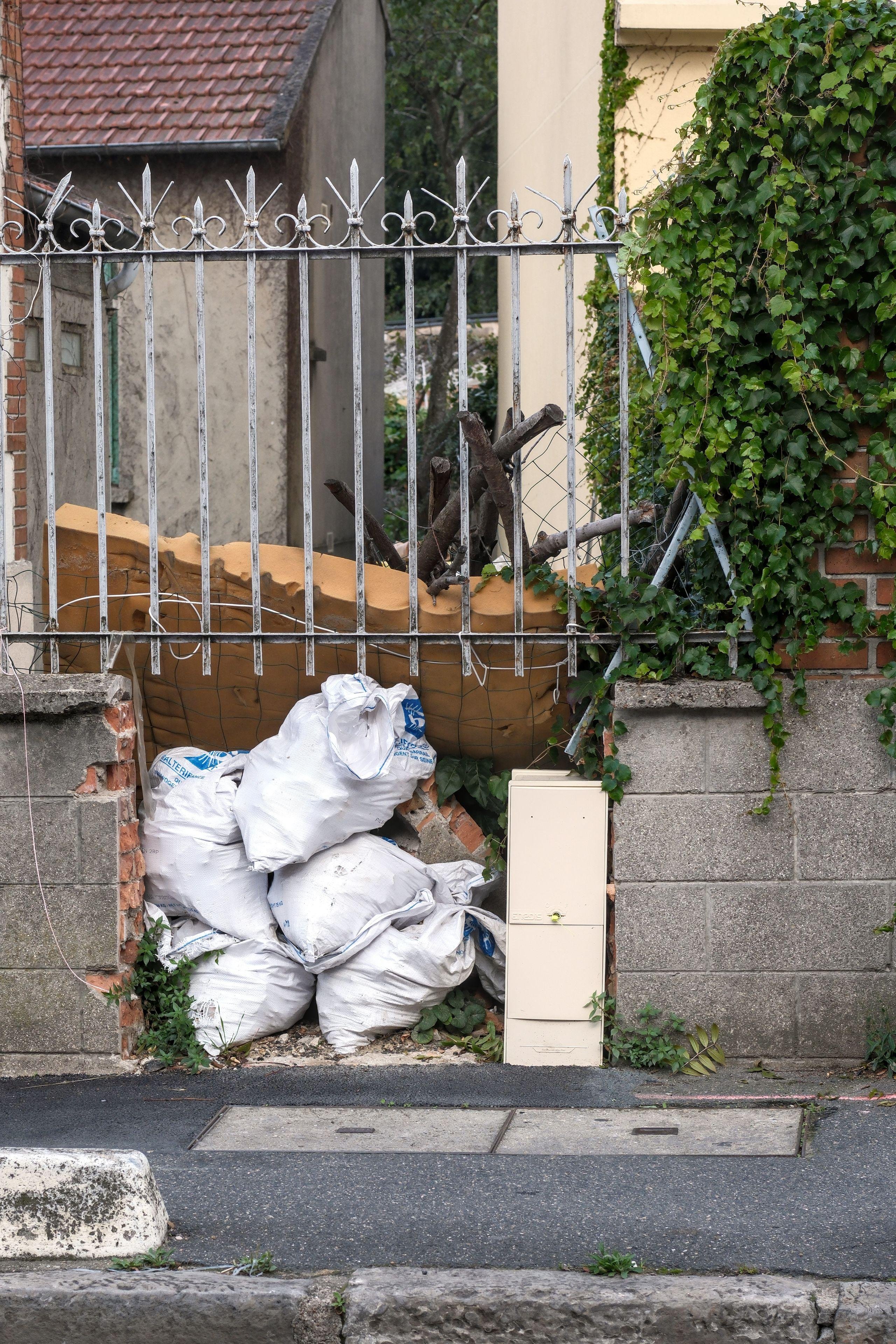 Photo verticale. Une barrière sur un muret dont un pan est manquant et comblé avec des sacs remplis de gravats, puis un vieux matelat et des grosses branches d’arbre appuyant sur le matelat