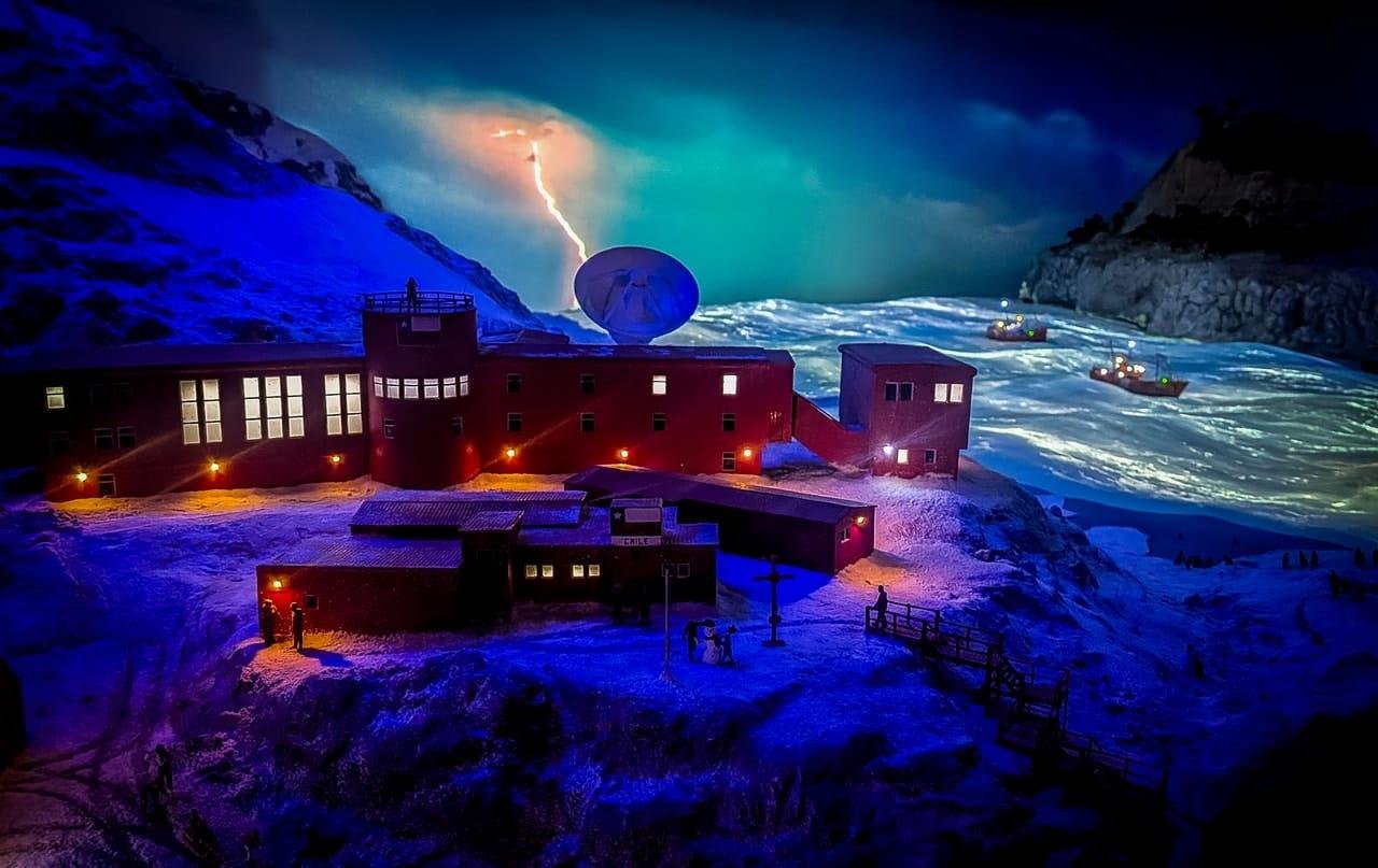 Snow-covered coastal research station at night with lit windows and orange exterior lights, a large satellite dish on the roof, and a lightning strike in a dark sky above an icy bay with small boats near rocky cliffs.
