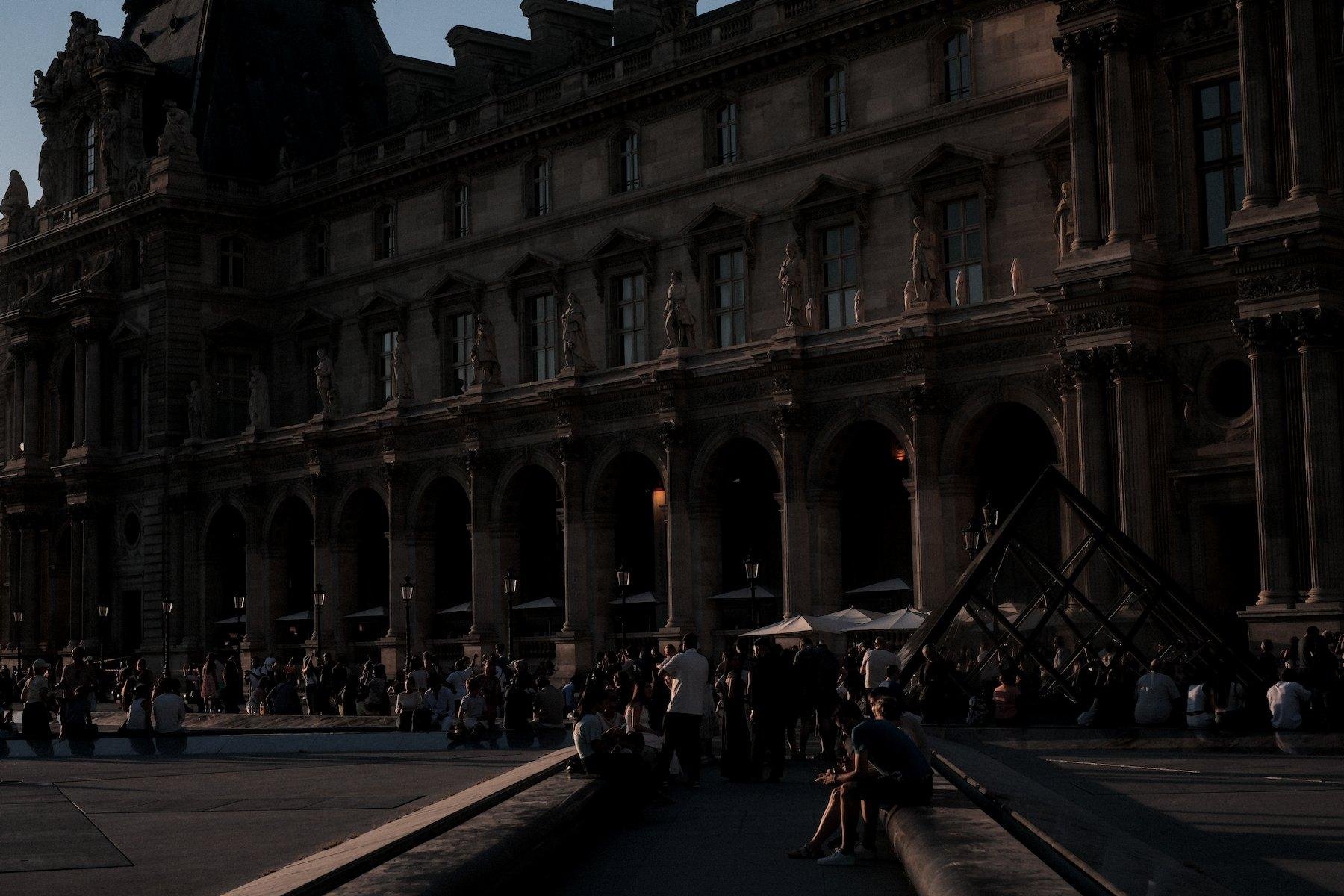 Photo horizontale. Sur le parvis du Louvre, plein de gens installé en train de regarder l’eclipse, dans une lumière tamisée (on est 3mn après de maximum)