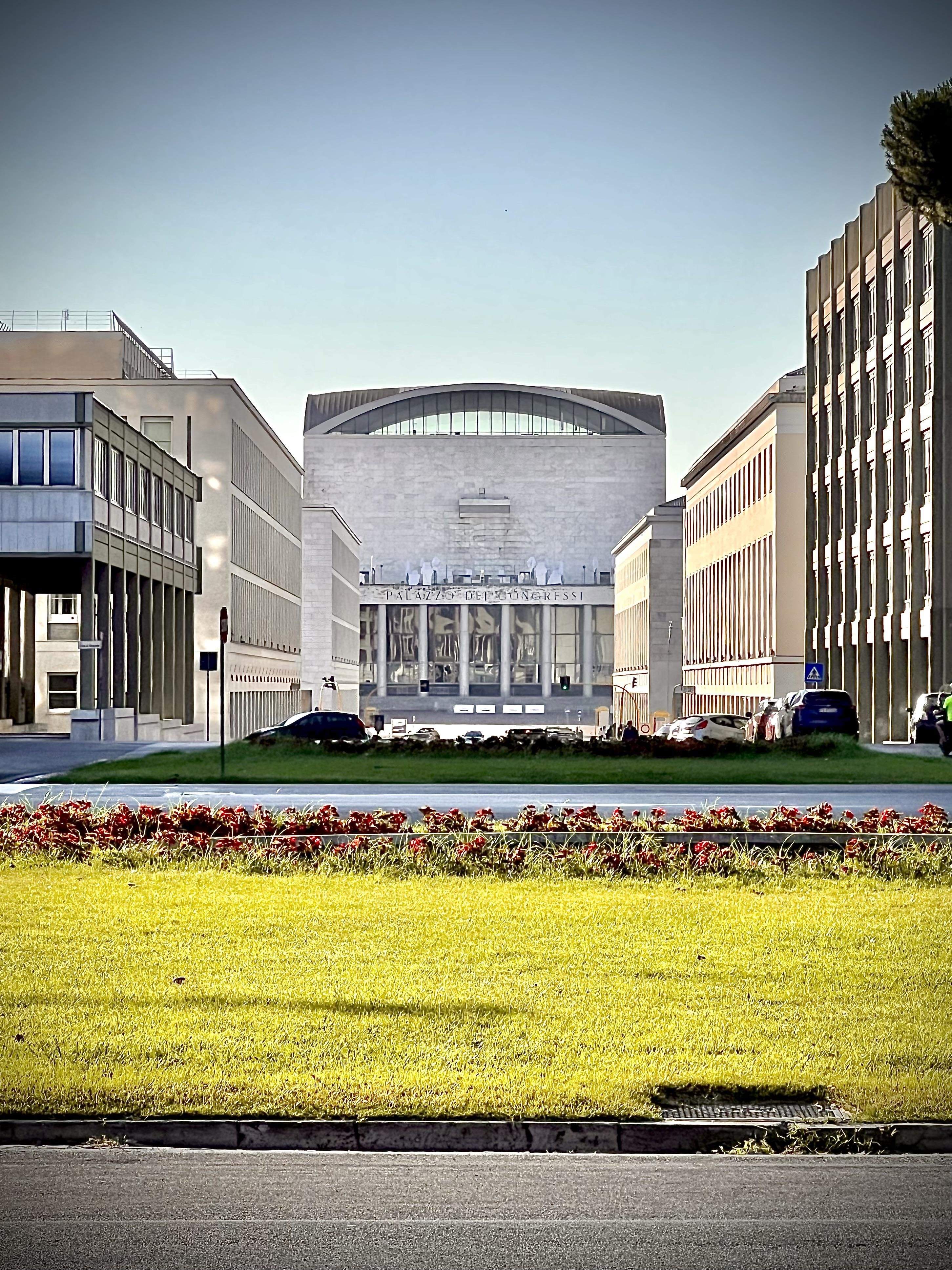Edificio denominato “Palazzo dei Congressi” con uno stile architettonico moderno. Ha una facciata simmetrica con grandi finestre di vetro. La struttura è affiancata da altri edifici. In primo piano, c’è un’area erbosa con un’aiuola di fiori rossi. L’ambientazione complessiva suggerisce un contesto urbano, possibilmente un distretto congressuale o governativo.