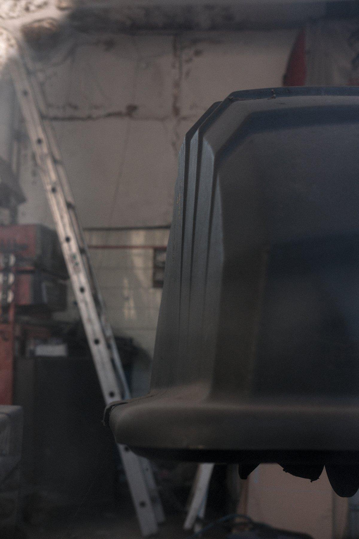 Photo verticale. Au premier plan, une espèce de couvercle en plastique noir faisant penser à un demi pneu de gros camion (mais ce n’est pas ça). En arrière plan, une grand échelle appuyée contre un mur. Avec plein d’étagères
