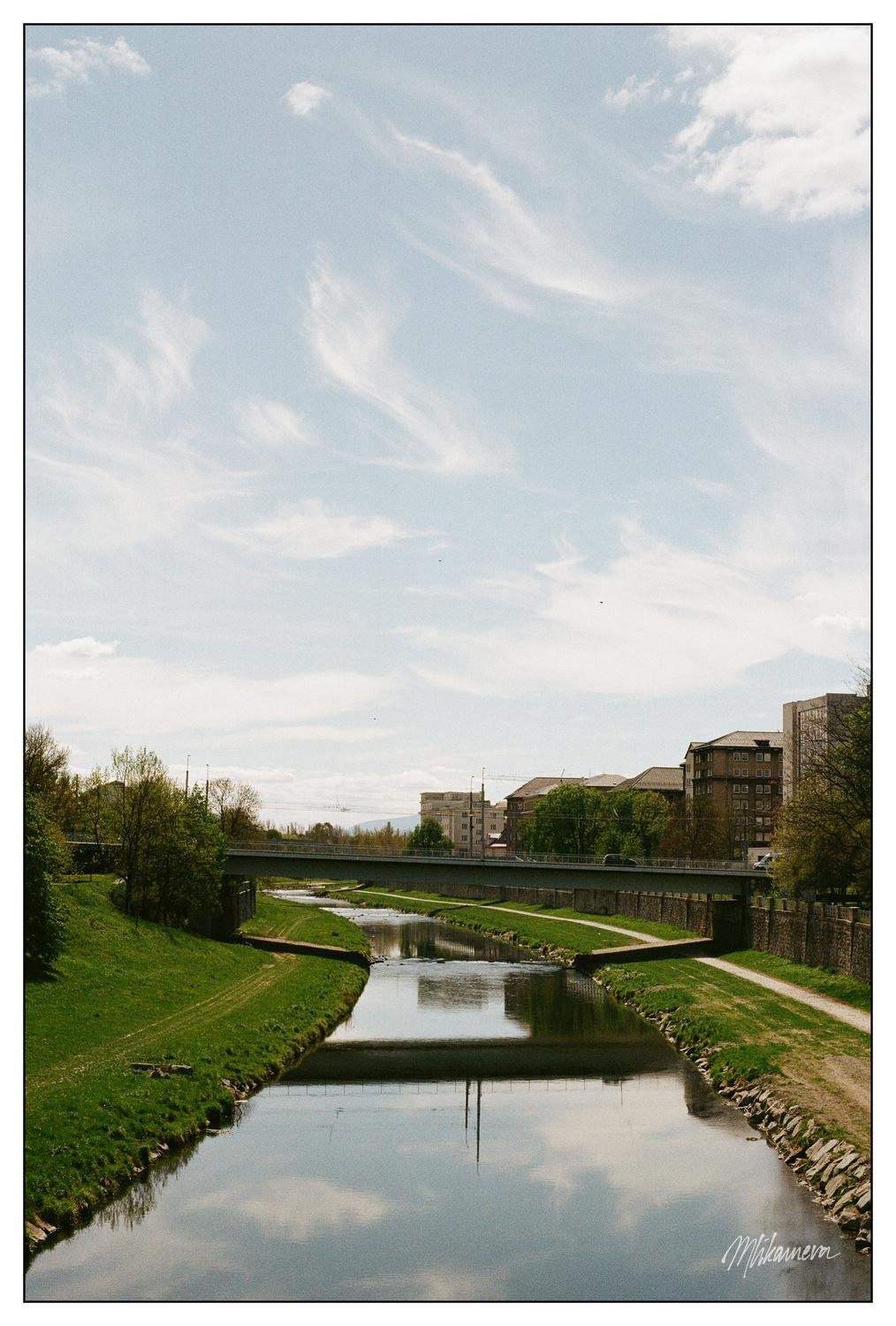 River with grassy banks flowing through an urban area, bordered by trees and buildings. Two bridges cross over the river. The sky is mostly clear with a few wispy clouds. Reflections of the surroundings are visible on the river's surface.