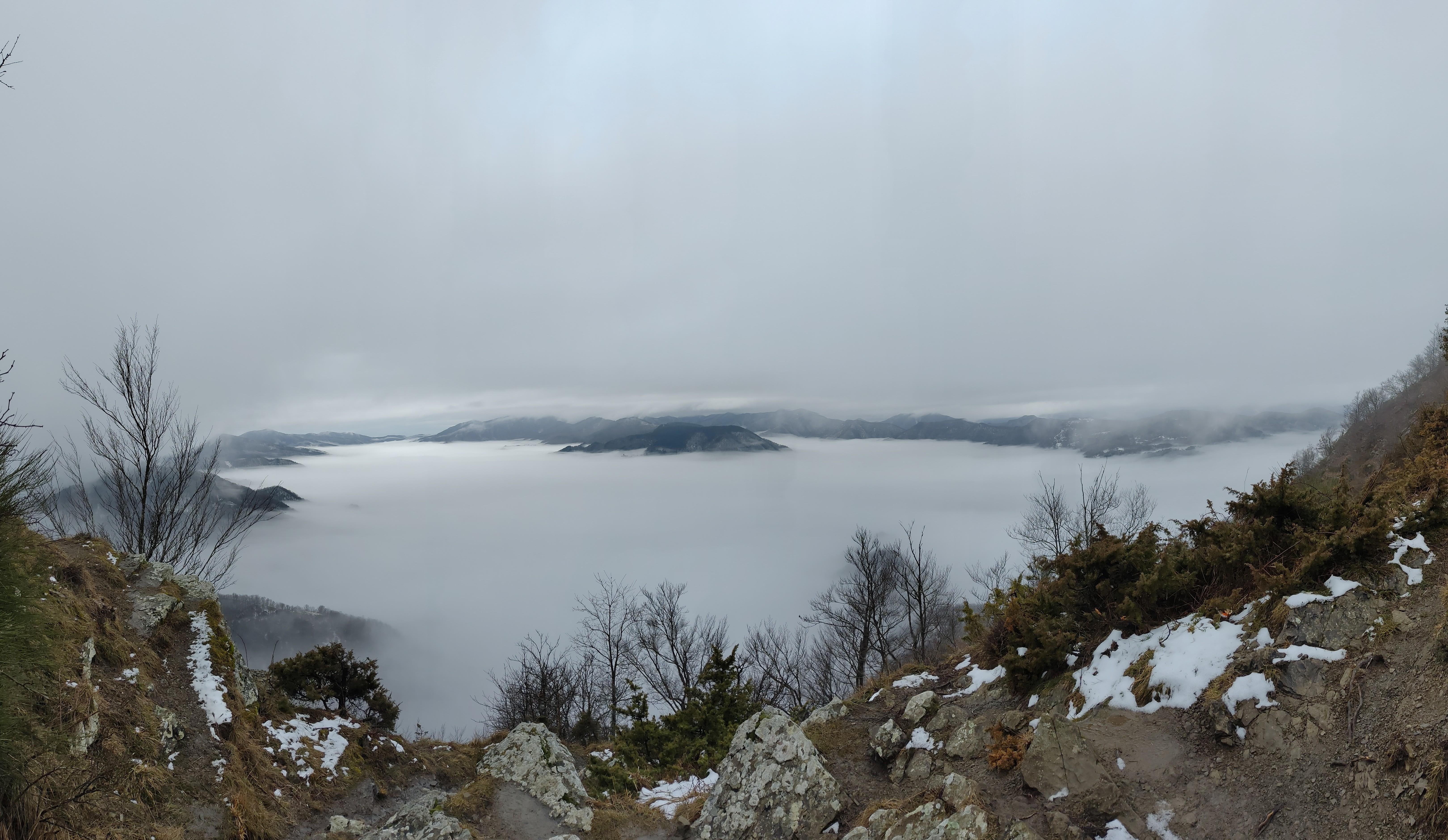 The image shows a panoramic view of a foggy valley from a high vantage point. The landscape is covered with dense fog, creating a sea of white that engulfs the hills and parts of the forested area. Bare trees and some patches of snow are visible in the foreground, with rocky terrain on either side. This perspective from the hillside or mountain overlooks the foggy valley, giving a serene and mysterious atmosphere.