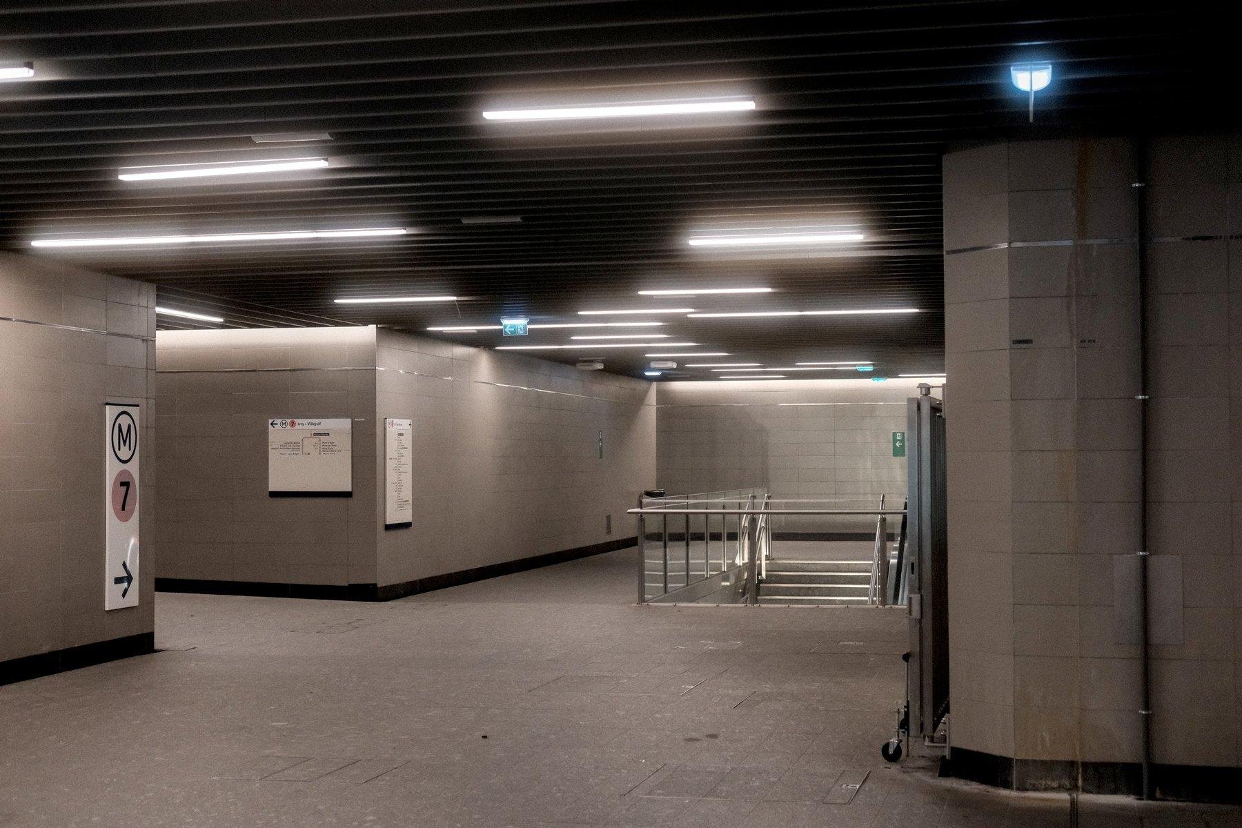 Photo horizontale. Un couloir du métro qui par à gauche et tout droit vers des escalier. Personne n’est en vue