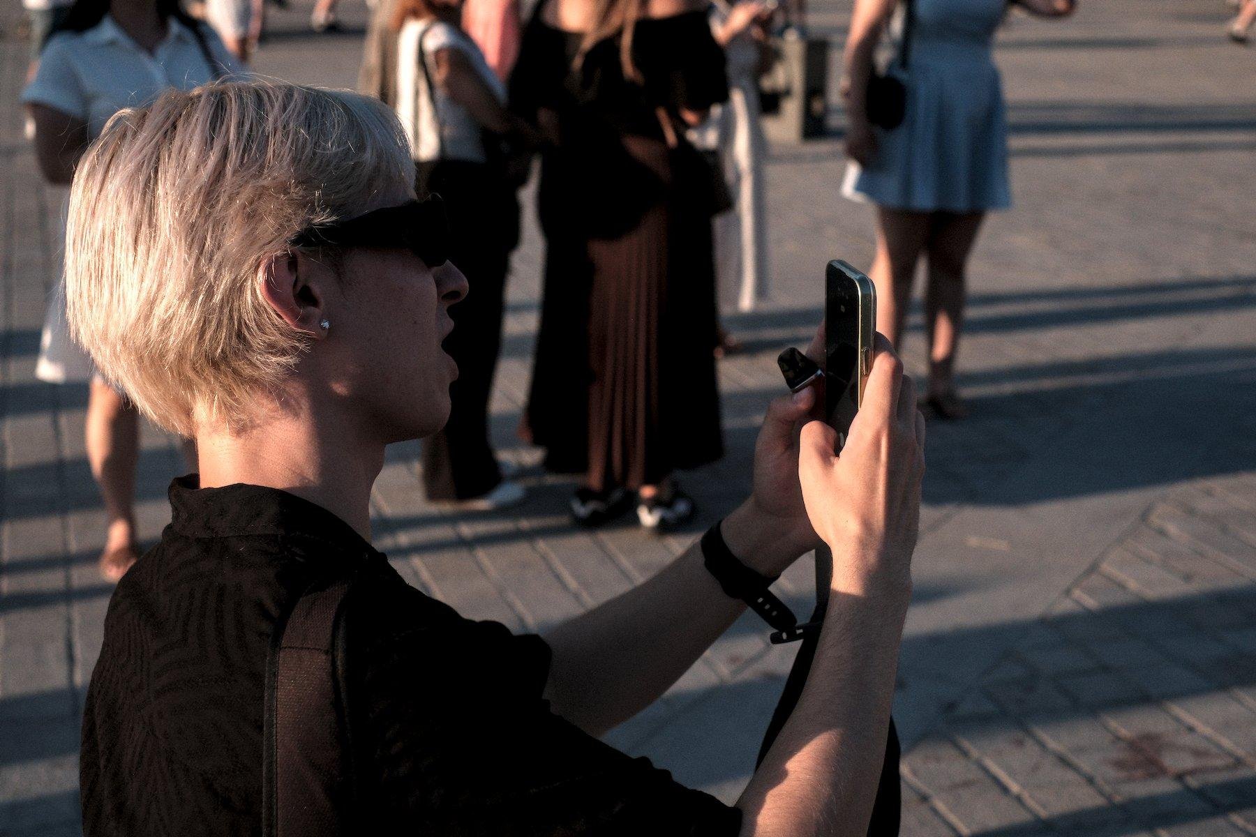 Photo horizontale. Portrait de profile (le droit) d’un homme blond, lunettes de soleil, haut noir. Il est entrain de prendre une photo avec un téléphone. Du monde en arrière plan, flous
