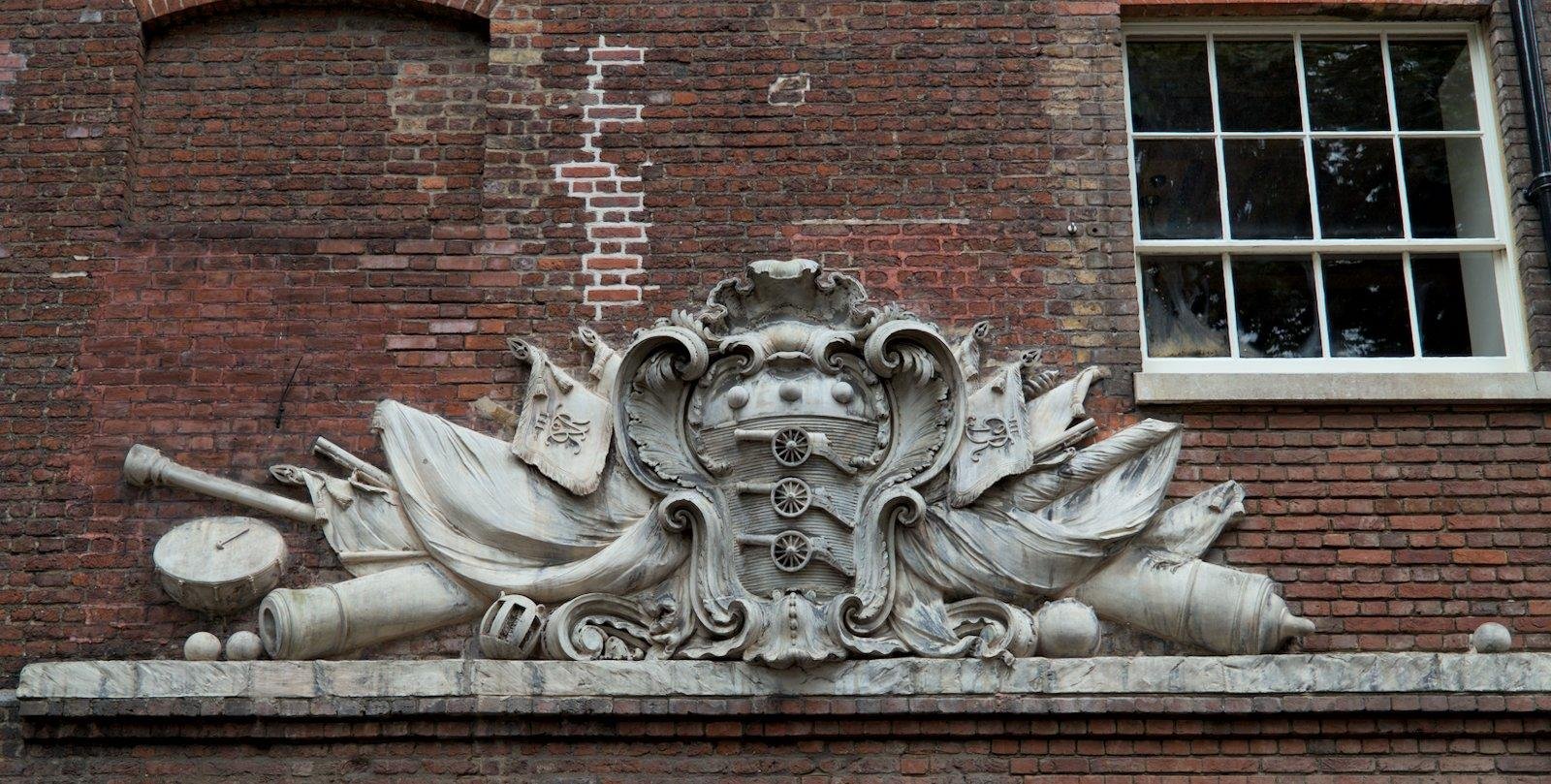 Photo format paysage, montrant un bas relief avec des armoiries royales, sur un mur de brique. On voit aussi une fenêtre quadrillée à droite