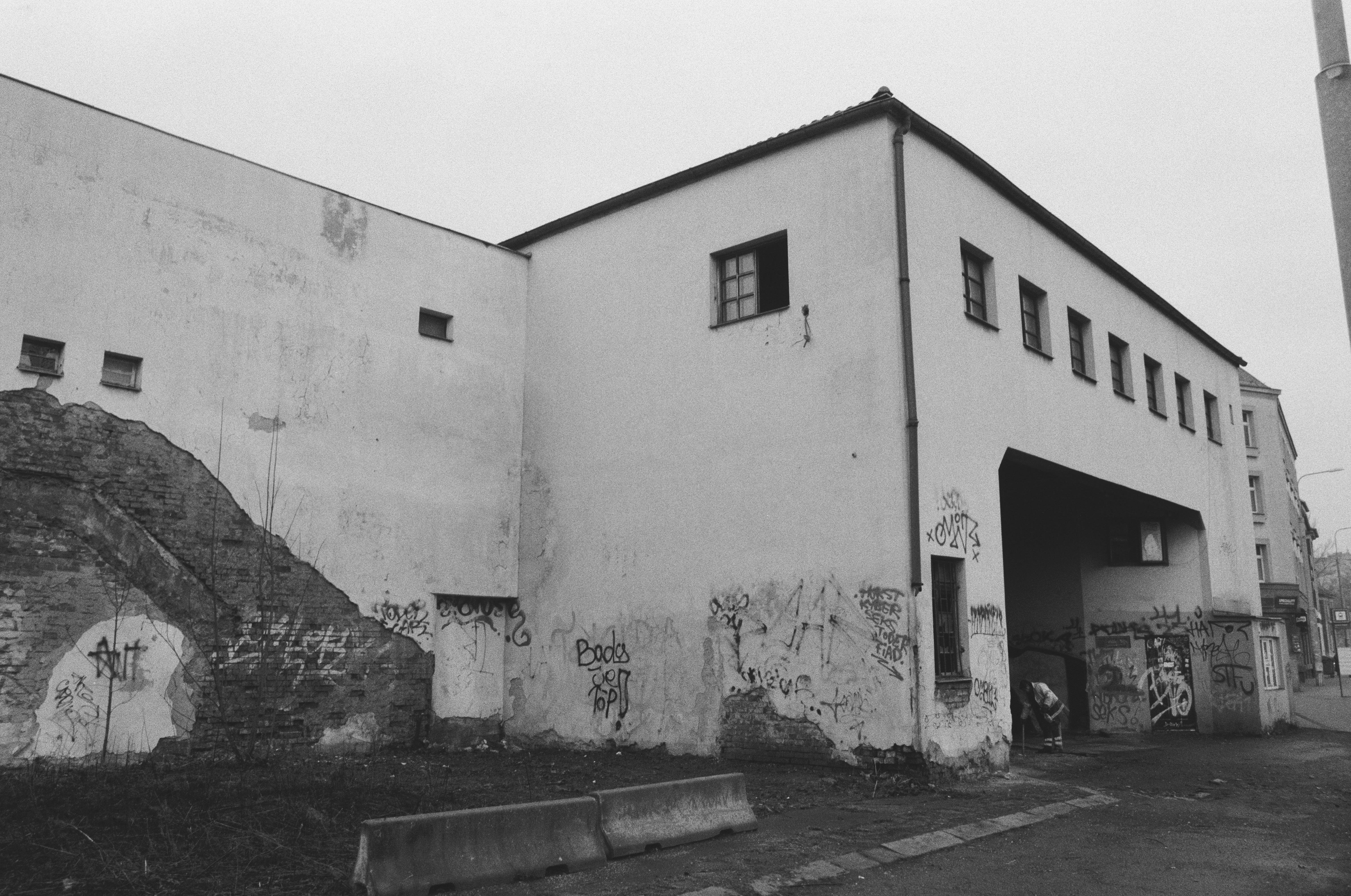 The image depicts a black-and-white photograph of an old building with visible graffiti on its walls. The structure appears worn and neglected, with parts of the brickwork exposed and rough terrain around it. The scene has an urban, somewhat desolate feel.