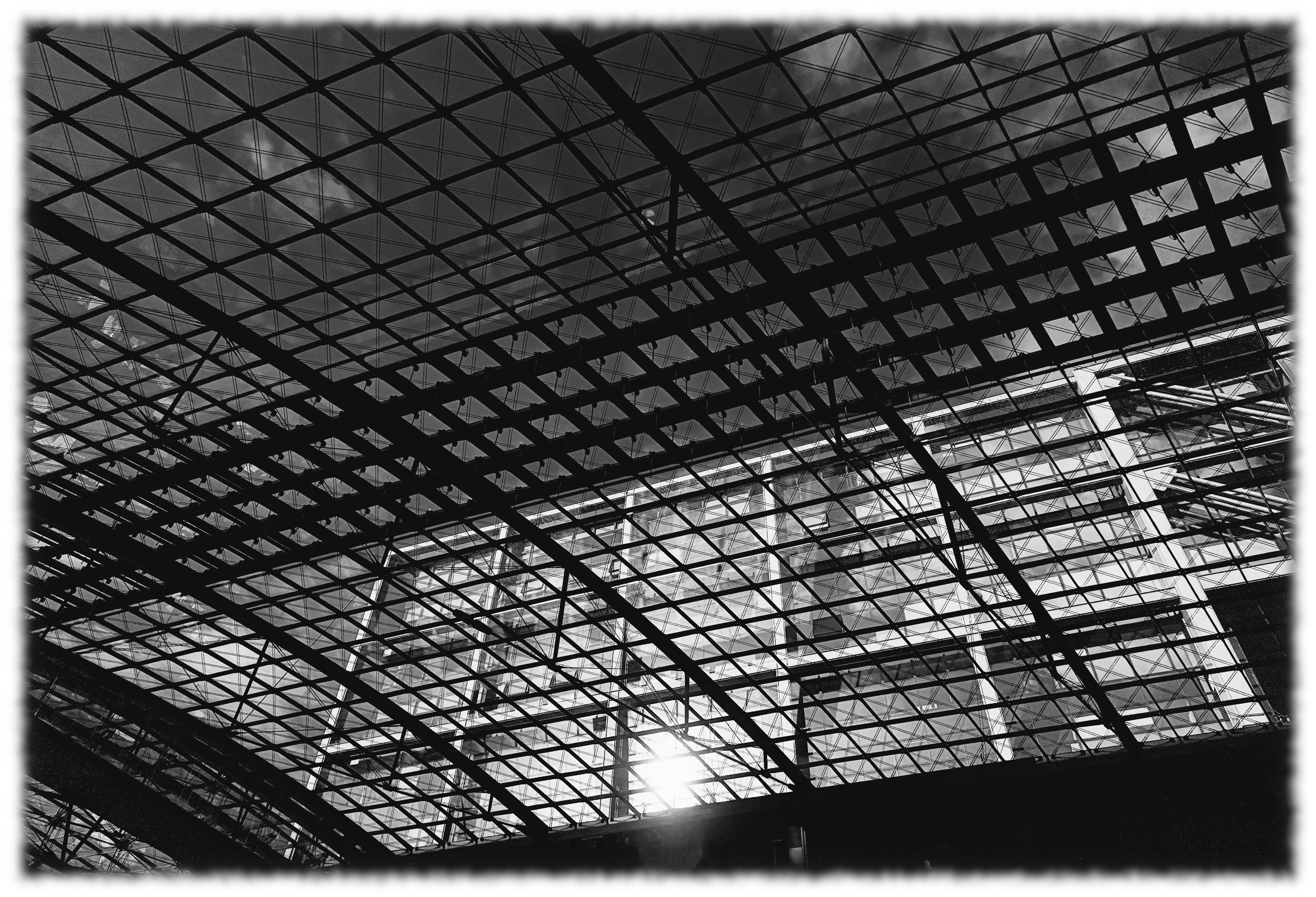 Upward view of a large arched glass-and-steel roof with a crisscross grid of beams, sunlight shining through near the lower left and a partial building facade visible at the right edge.