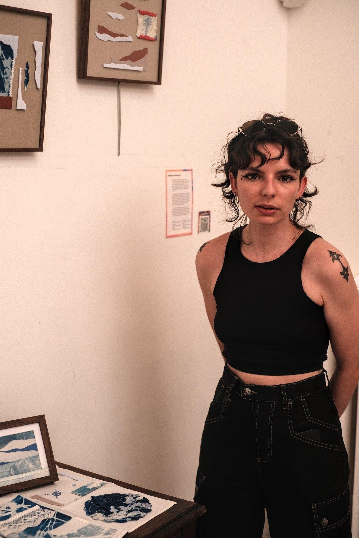 Photo verticale. Portrait d’une jeune femme brune au cheveux attachés, en débardeur noir et pantalon noir, les bras dans le dos, regardant la caméra en parlant de ses œuvres