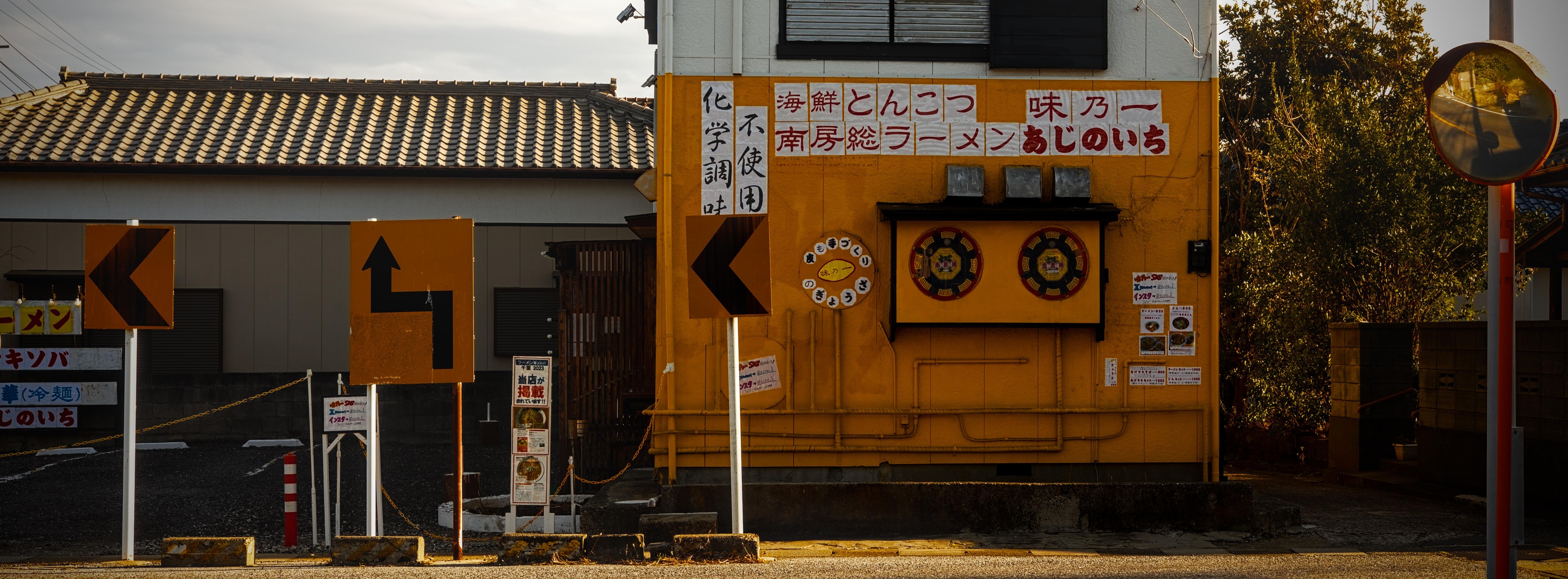 The facade of a two-story ramen restaurant. The ground floor is painted in a yellow-orange tone that is similar to the road signage in front of it. The building itself is covered in inscribings with the menu, social media accounts, etc. A view mirror is visible to the extreme right of the picture.