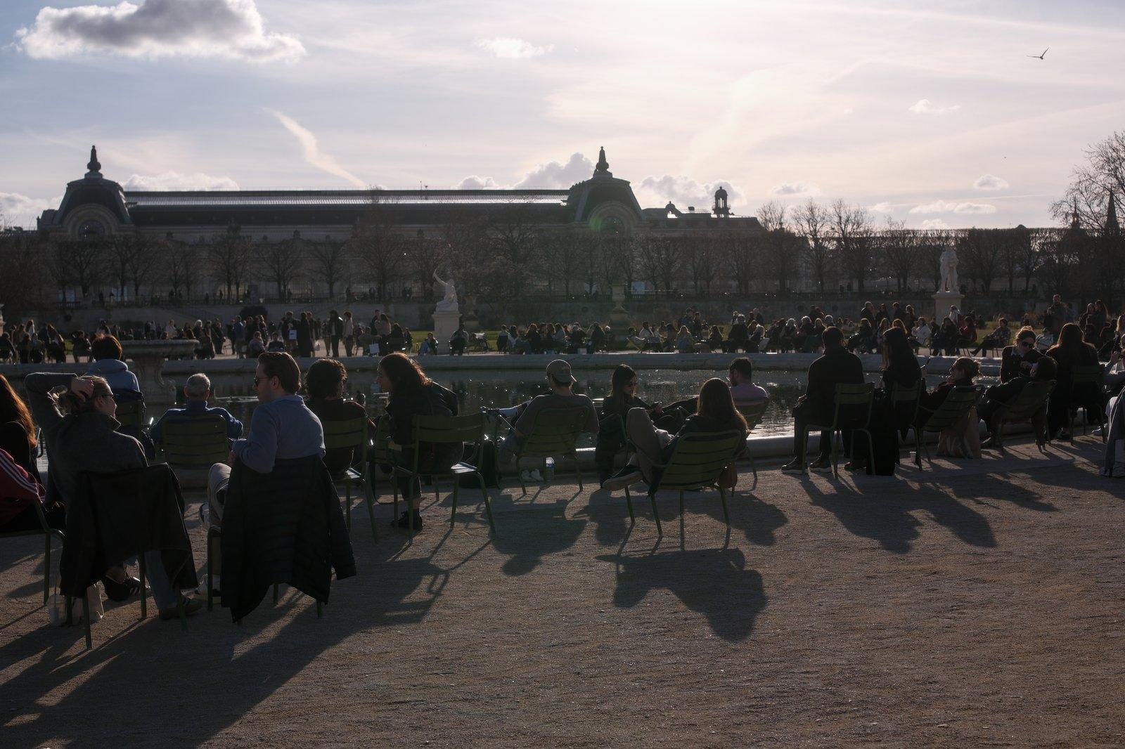 Photo horizontale. Plein de gens assis sur les chaises métaliques vertes du jardin du Luxembourg à Paris, autour du lac artificiel, le Soleil les réchauffant et projetant leur ombres au sol