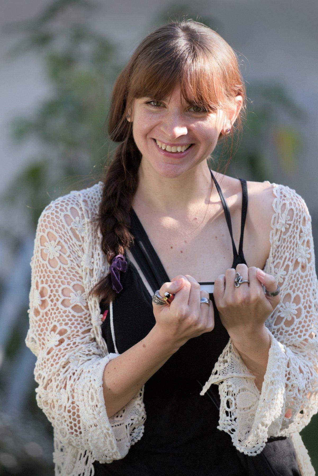 Photo format portrait d’une jeune femme brune aux cheveux longs coiffés en une natte qui tombe devant son épaule droite. Elle est habillée d’un débardeur noir à fines bretelles et un gilet fait en crochet avec du fil blanc au motif fleuri. Elle tiens entre ses mains un bloc note par en dessous, tenant le haut du carnet, avec le bas qui repose contre son torse. Elle regarde la caméra avec un grand sourire sympathique