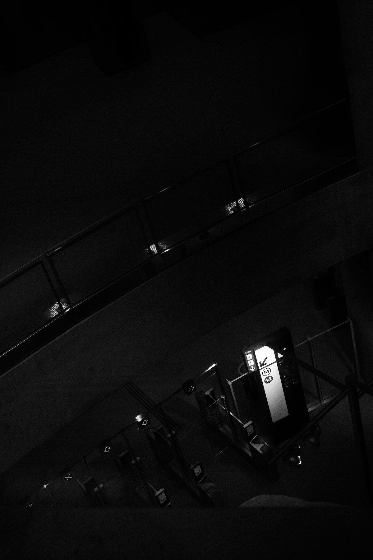 Photo verticale en noir et blanc. Plongée vers les bornes de validation de métro. Tout est très sombre sauf le panneau indiquant d’une flèche, la ligne 14