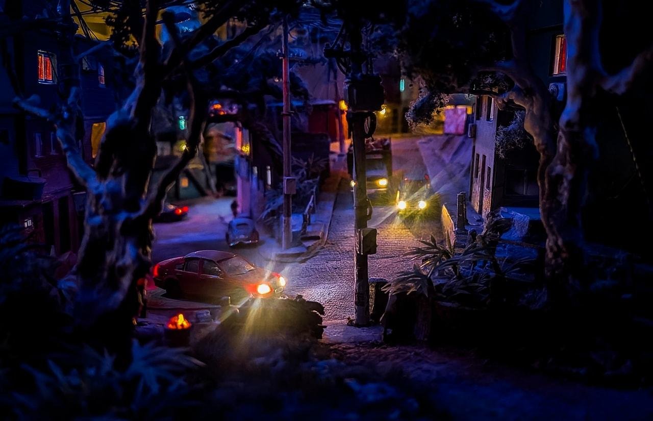 Nighttime miniature city street viewed through dark tree branches, with a cobblestone road between buildings, glowing streetlights, and several cars with headlights and taillights illuminating the scene.