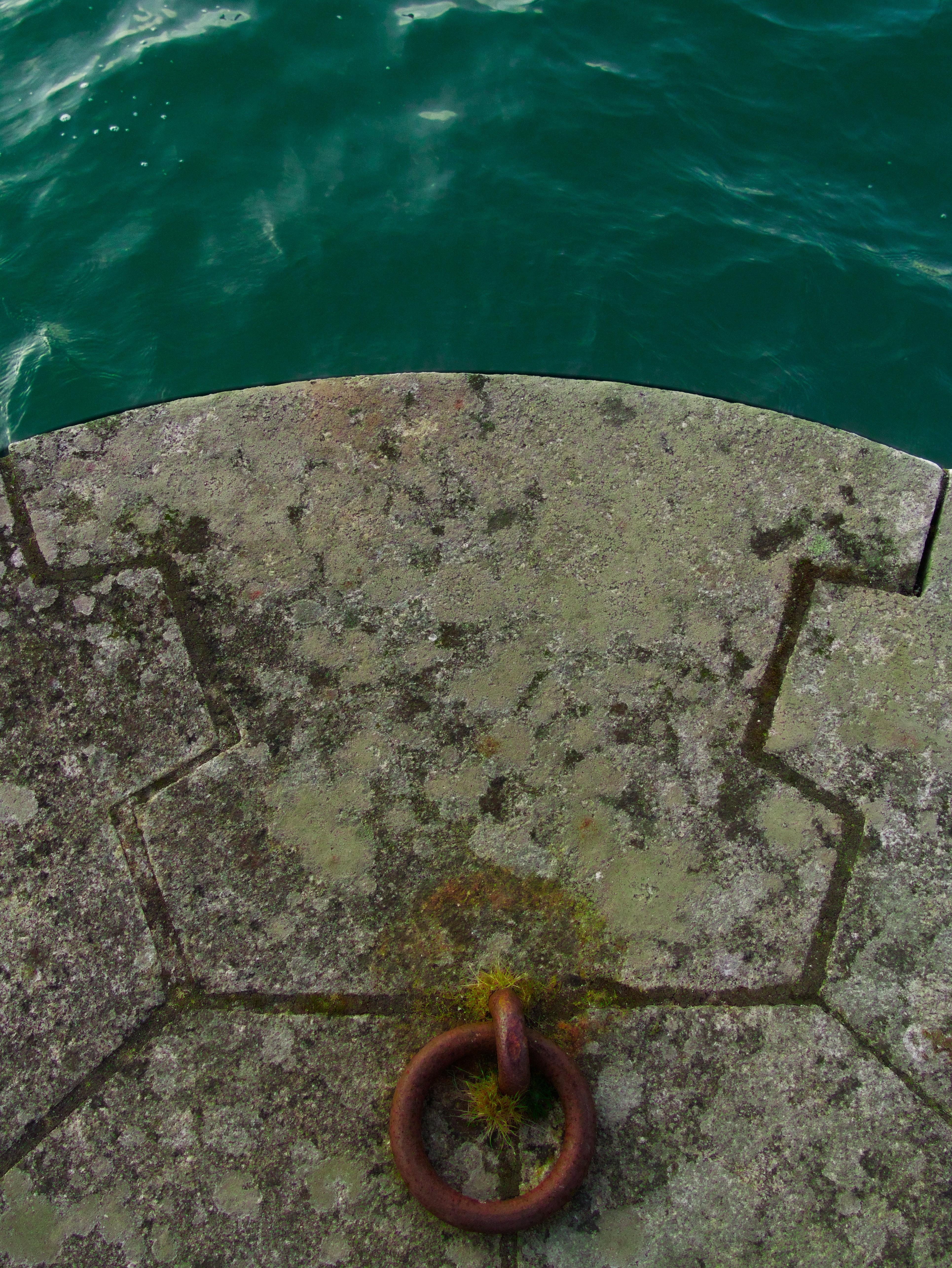 Weathered stone dock edge above green-blue water, with a rusted metal mooring ring embedded near the bottom center.