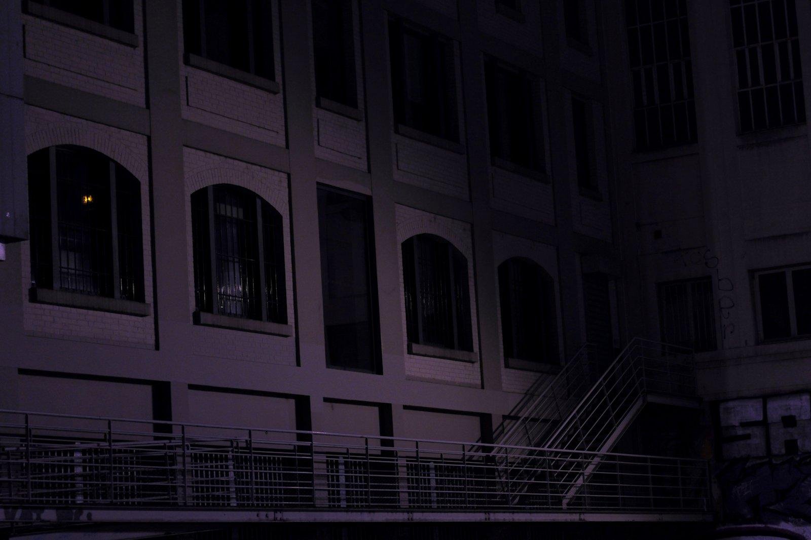 Photo format paysage, de nuit, de teinte violette, montrant un mur d’immeuble avec des fenêtres protégée de barreaux. Une rampe de passage en bas de l’image, qui mène à un escalier extérieur qui monte vers une porte presque invisible dans le noir