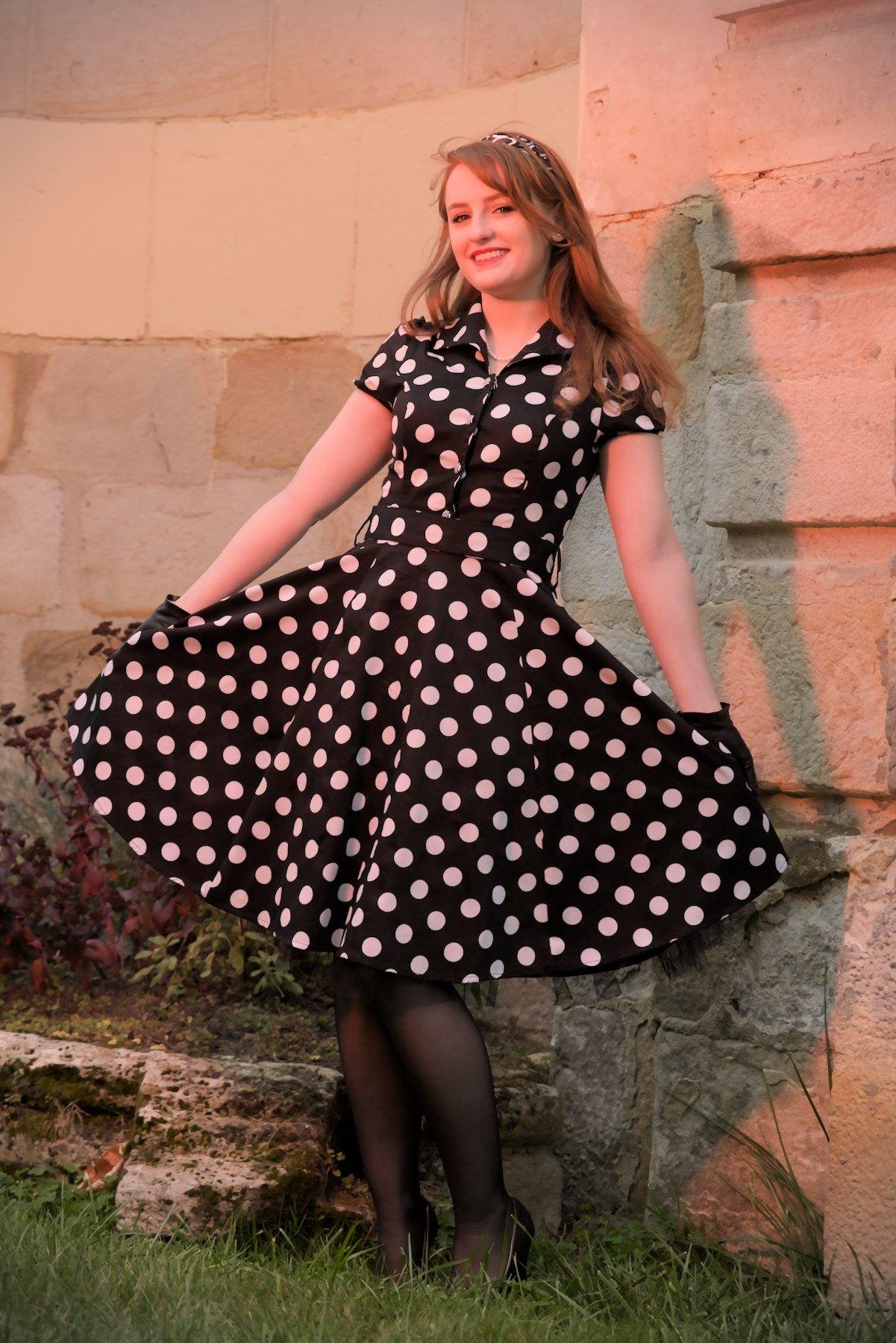 Photo verticale. Une jeune femme aux cheveux long roux, posant devant un mur de vieilles pierres, portant une robe noire à pois blanc, des collants noirs, des chaussures noires avec un petit nœud, des gants  en cuir noir et un serre tête noir avec des motifs blancs. Elle tiens sa robe pour l’écarter et la présenté à la caméra en souriant