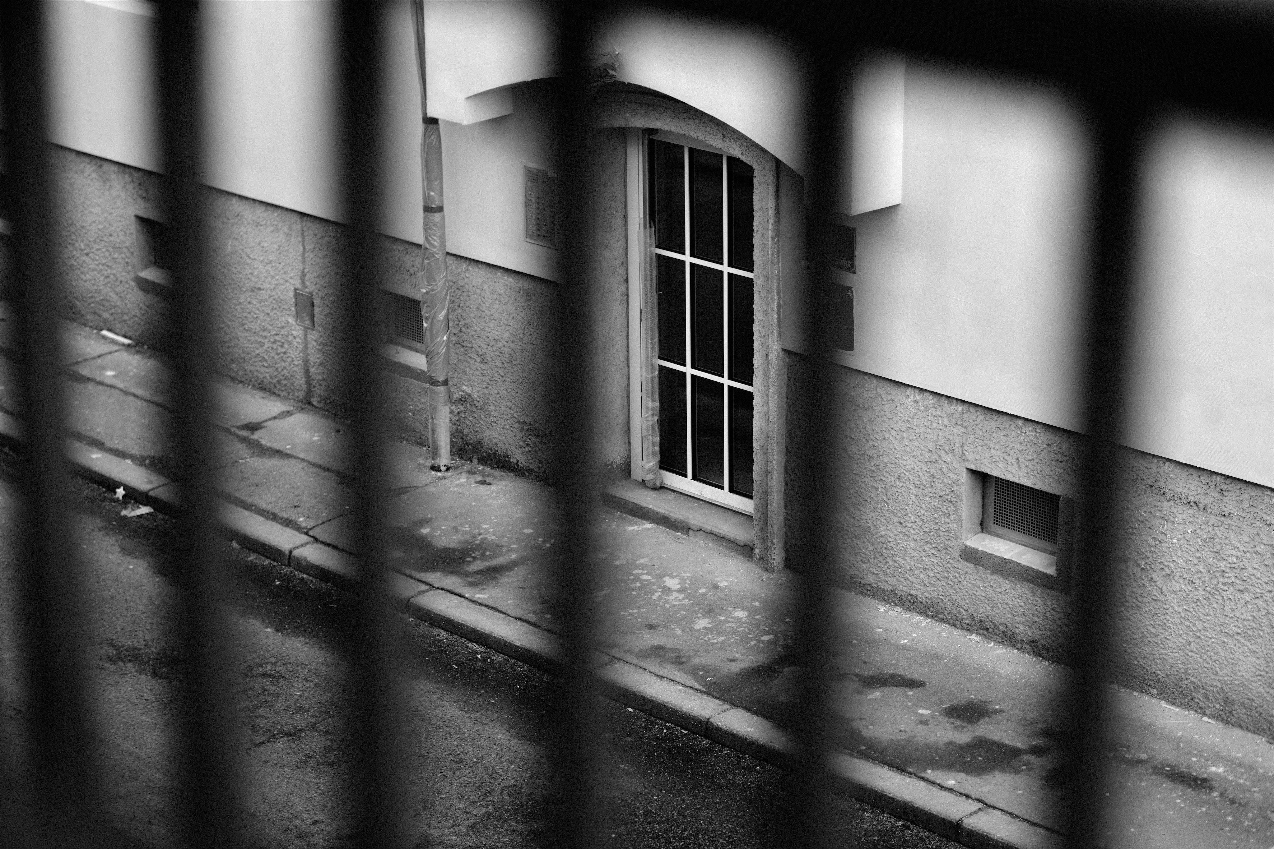 Schwarz-Weiß-Straßenansicht durch unscharfe vertikale Zaun, die einen Bürgersteig und einen Bordstein neben einer Gebäudewand mit einer gewölbten Tür mit einer vergitterten Glastür und einem kleinen rechteckigen Fenster auf der rechten Seite zeigen.