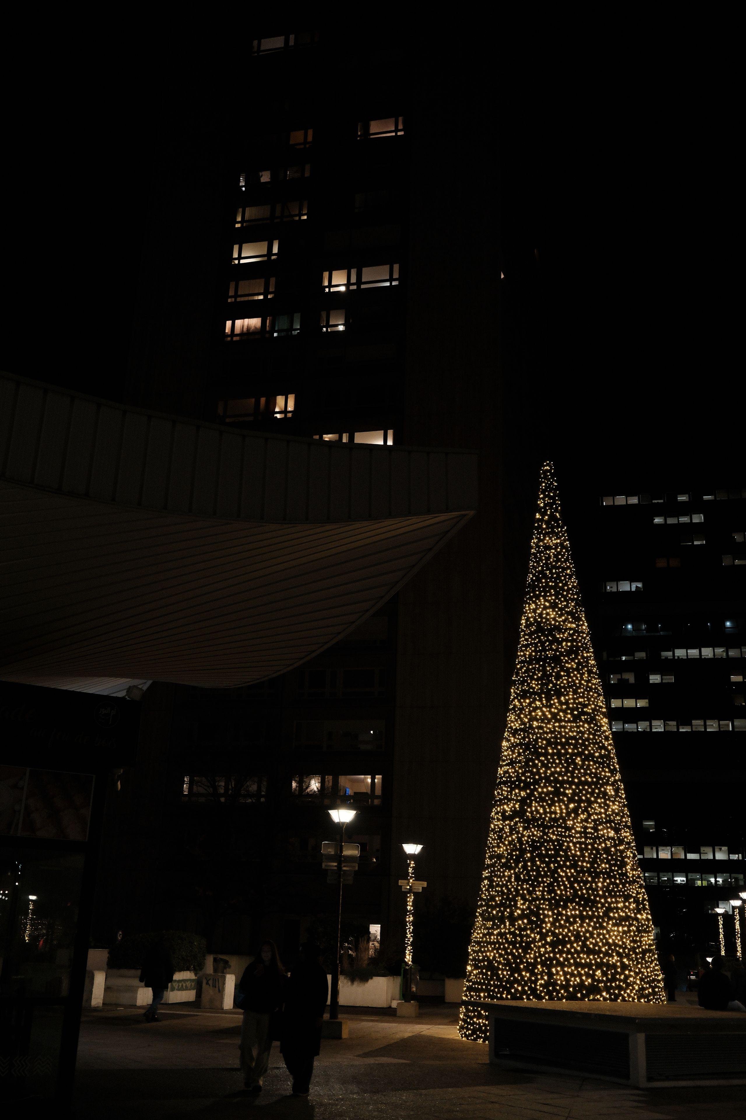 Photo verticale. Un sapin de noël stylisé, fait d’un cone de LED sur fond de hauts immeubles dont quelques fenêtres sont allumées.