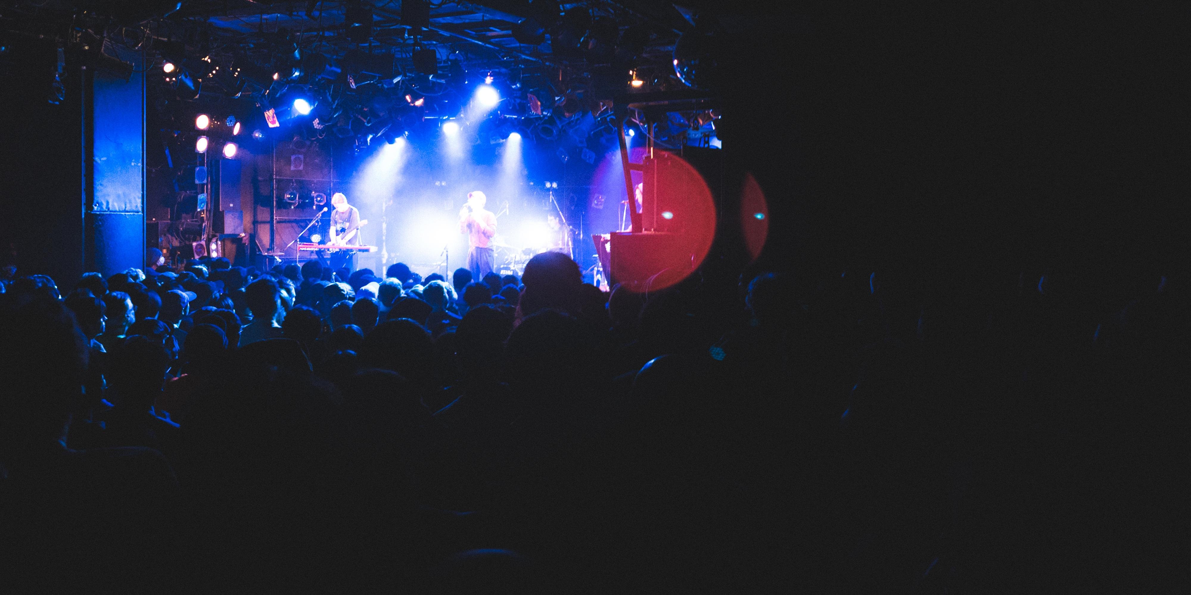 Concert scene with a densely packed audience in a dimly lit venue. Blue spotlights illuminate a stage where two performers stand with microphones, surrounded by musical equipment. The crowd is silhouetted in the foreground, creating a contrast with the bright lights on stage.