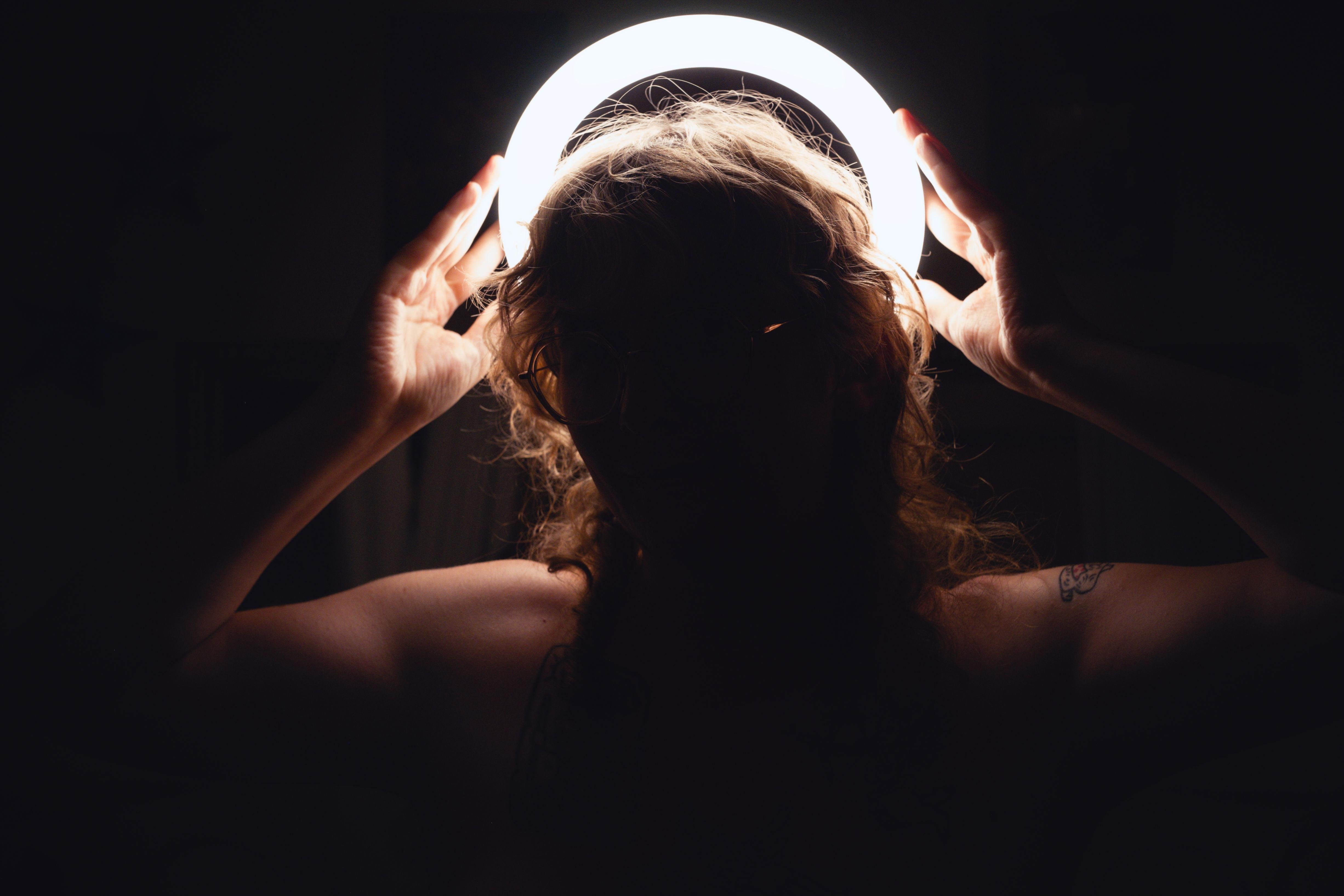 Autoportrait pris avec un anneau lumineux porté derrière la tête. L'anneau éclaire mes cheveux bouclés, mes mains et mes épaules. Le reste est dans l'obscurité.