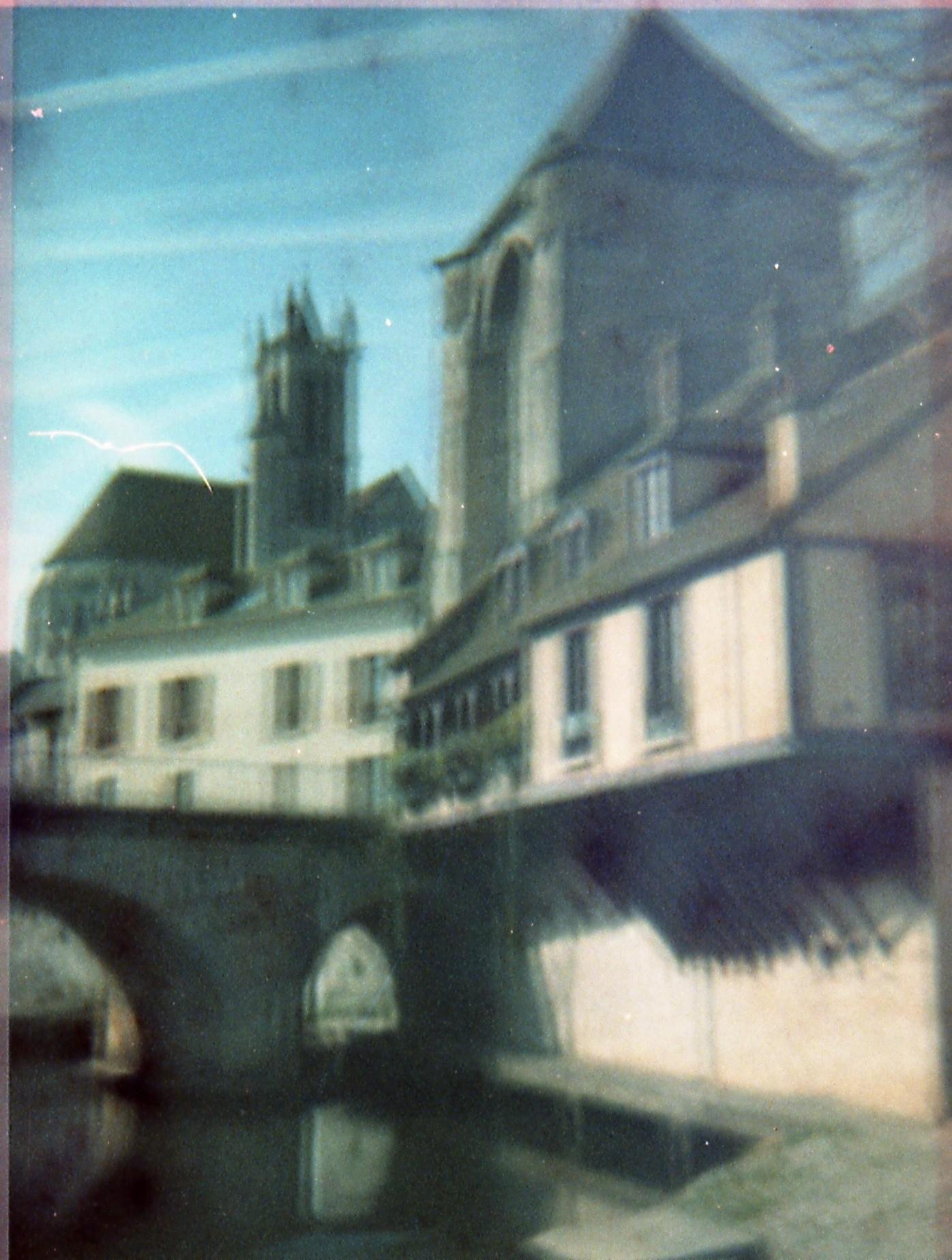 Blurred view of a riverside town with a stone church and tall tower behind a row of buildings; their reflections appear in the water in the foreground under a blue sky.