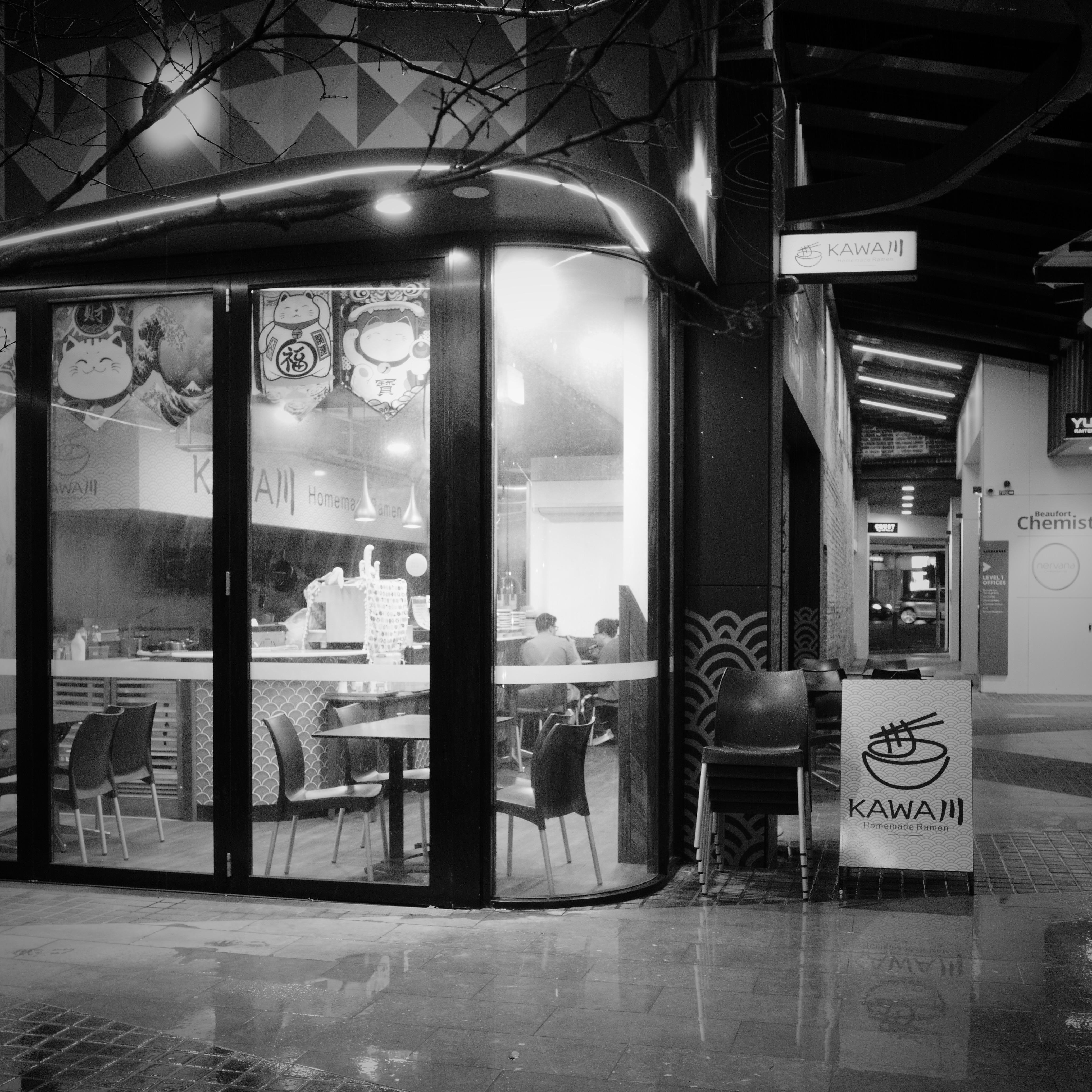 Black-and-white street photo of a corner ramen shop with curved glass windows, “KAWAII” signage, and a sandwich-board menu by the entrance; empty chairs and tables visible inside, with wet pavement reflecting the storefront.