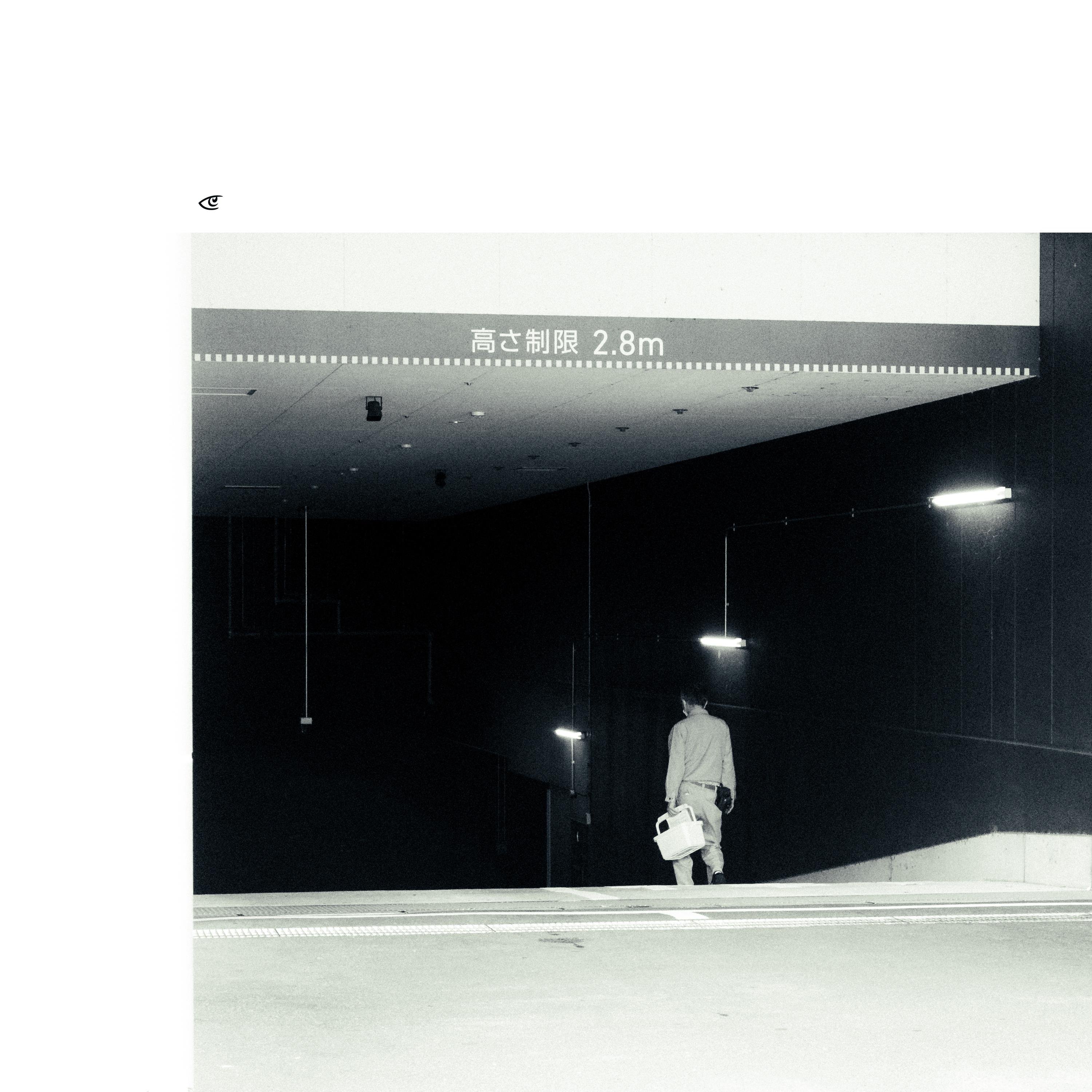 Black-and-white photo of a person in light clothing walking down a sloped ramp into a dark underground parking entrance, carrying a white bag; overhead sign reads “高さ制限 2.8m” with fluorescent lights along the ceiling.