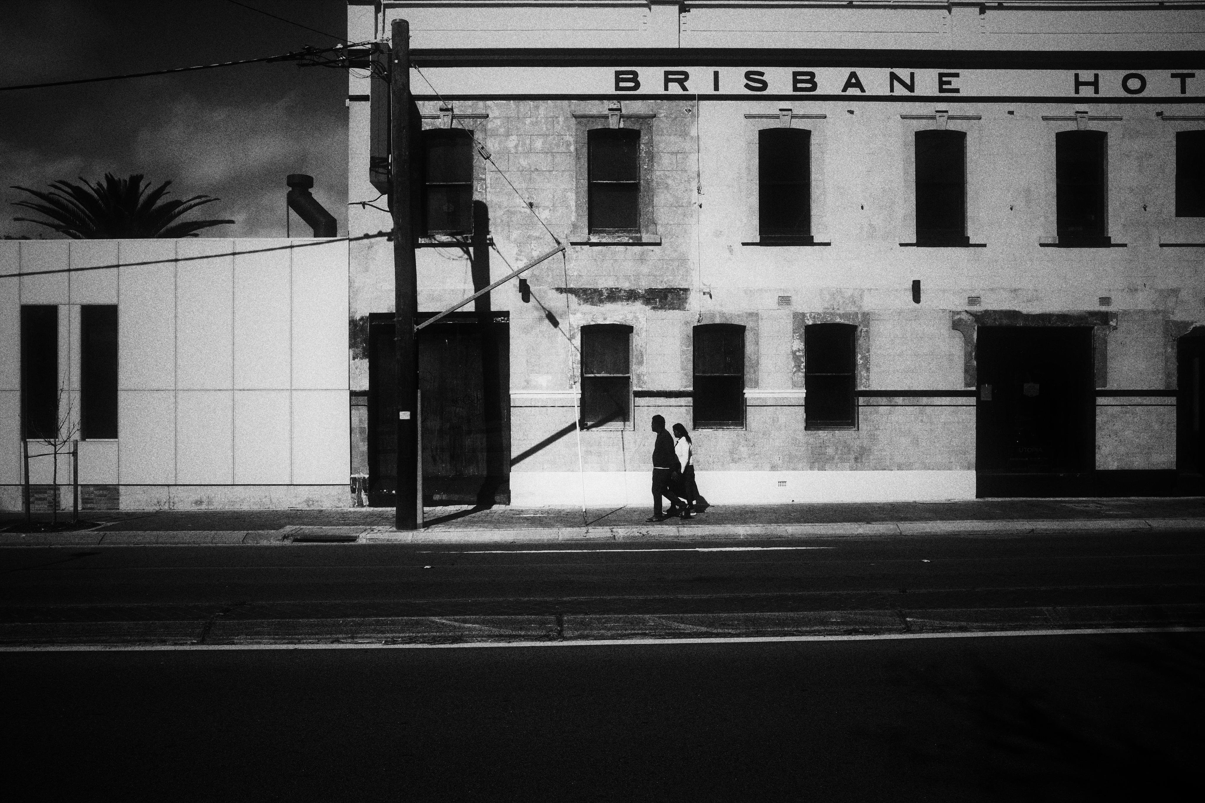 Black-and-white street scene with two pedestrians walking along a sidewalk in front of a weathered two-story building labeled “BRISBANE HOT(EL),” featuring multiple dark windows, a large doorway, power poles and overhead wires, and an empty road in the foreground.