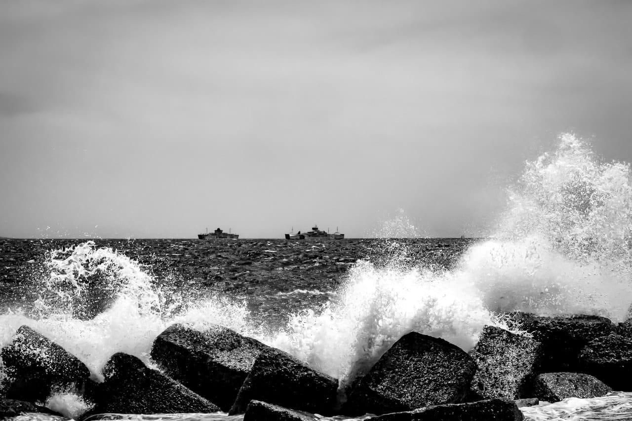 Black-and-white seascape with waves crashing against large rocks in the foreground, white spray rising, and several small boats on the horizon under a cloudy sky.