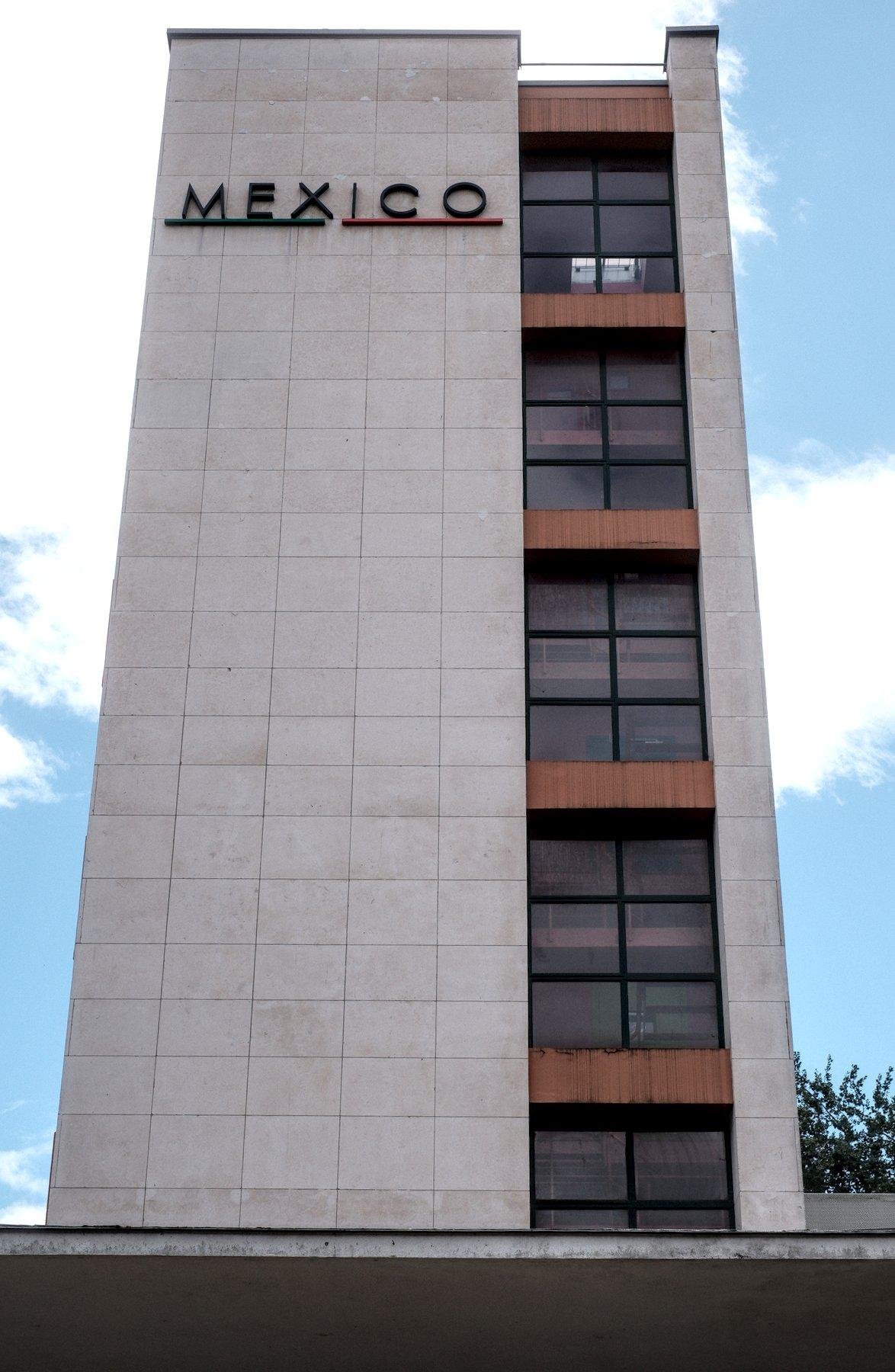 Photo verticale. Contre plongée d’un petit immeuble de 5 étages, au ⅘e fait de dalles blanche, et sur la droite, des fenêtres avec chaque plafond marqué d’une séparation orange. En haut de la partie blanche, Mexico est écrit et souligné aux couleurs du drapeau mexicain